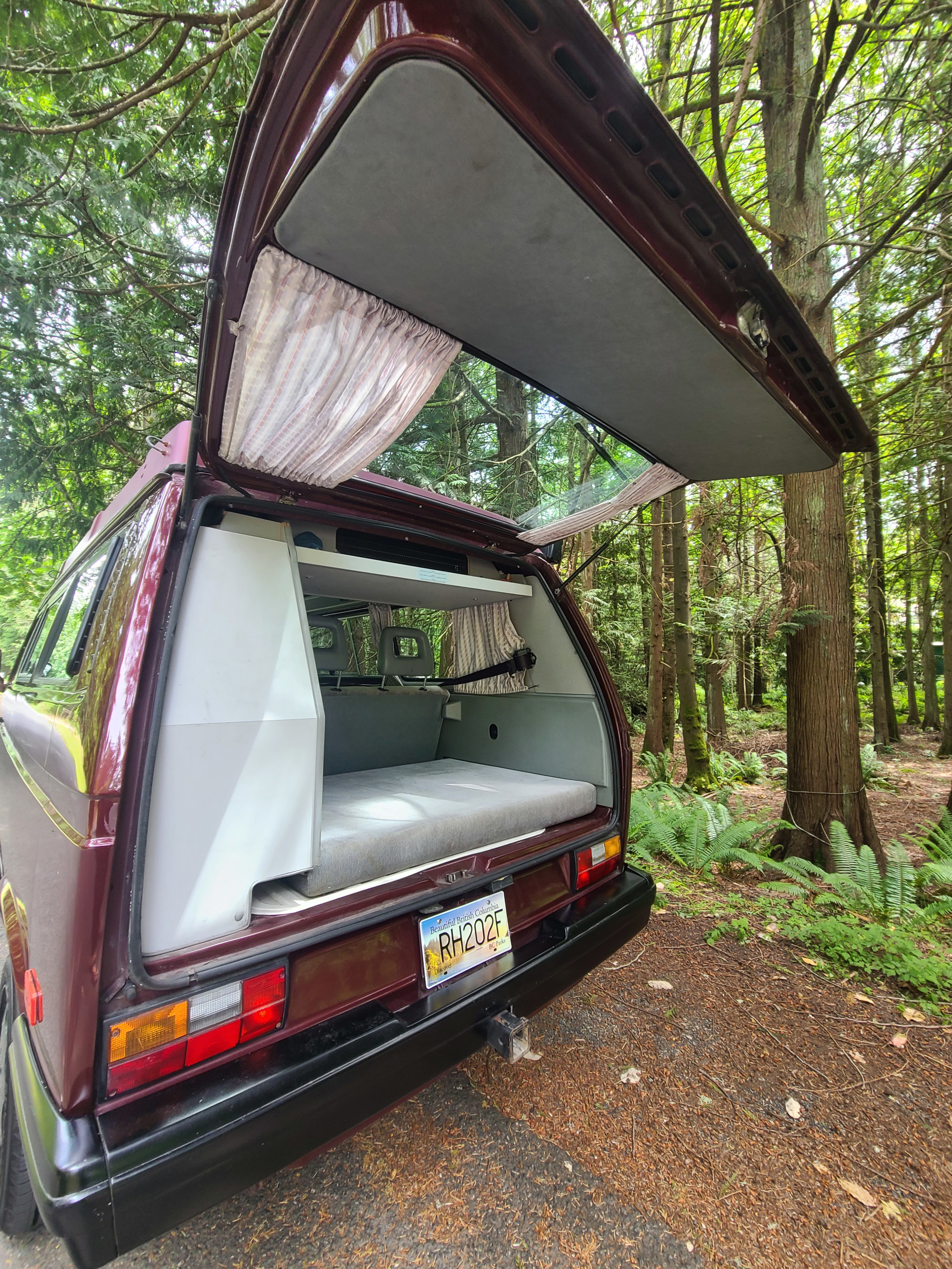 Maroon camper van, ideal for your next RV rental, with its rear hatch open revealing a cozy bed in a peaceful forest setting.. Volkswagen T3 Mulitvan 1991