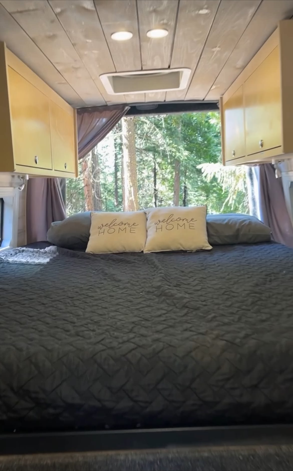 Comfortable RV rental interior with a cozy bed, "welcome HOME" pillows, and a serene forest view.. Mercedes Sprinter 2020