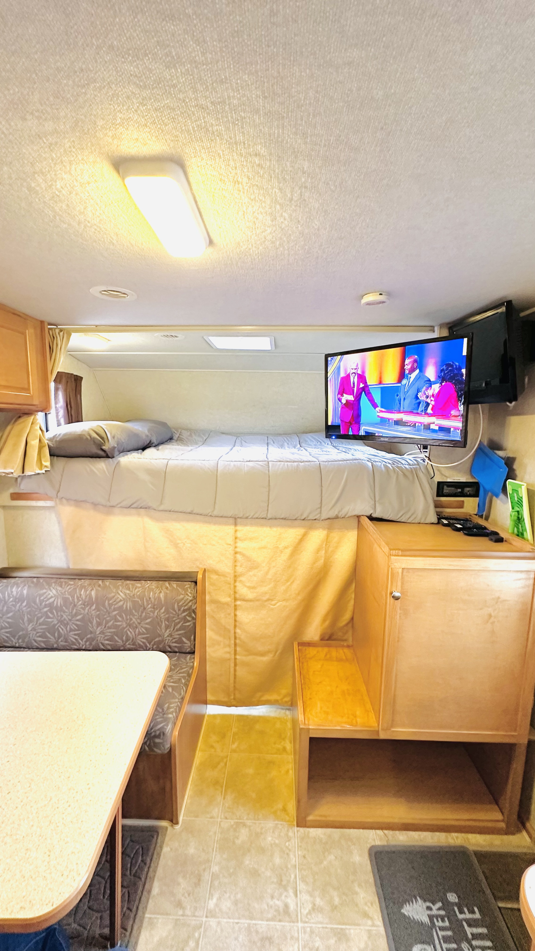 Cozy RV rental interior showing a raised sleeping area with a bed, a mounted TV playing a game show, and a dinette.. Winnebago Chalet 2009