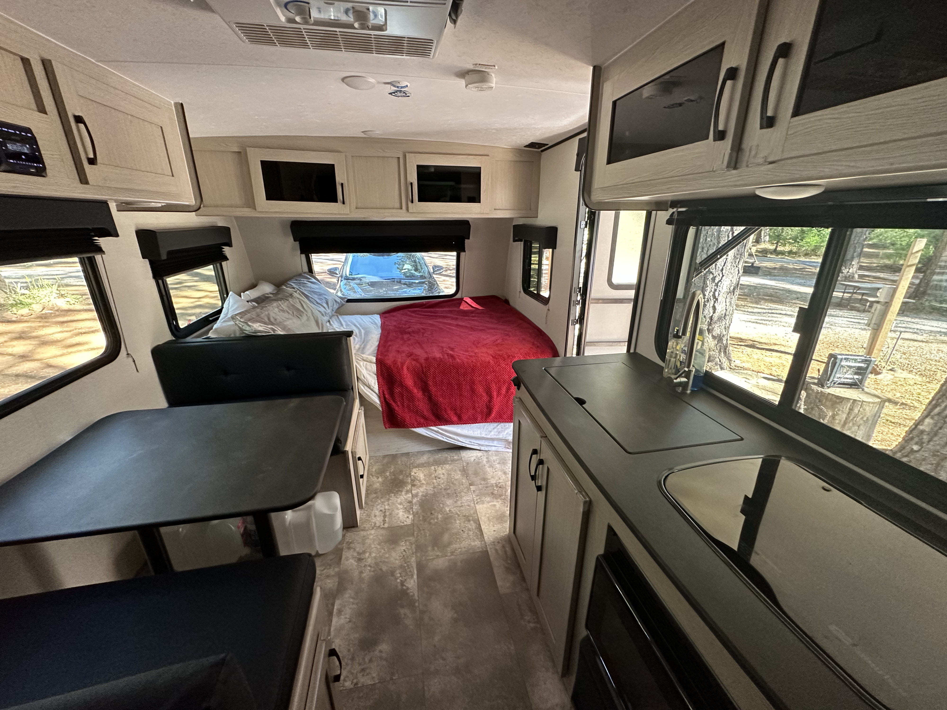 Modern RV rental interior with dinette (left), rear bed, and kitchen (right). Features light cabinets, black counters, and outdoor views.. Apex Nano 2023