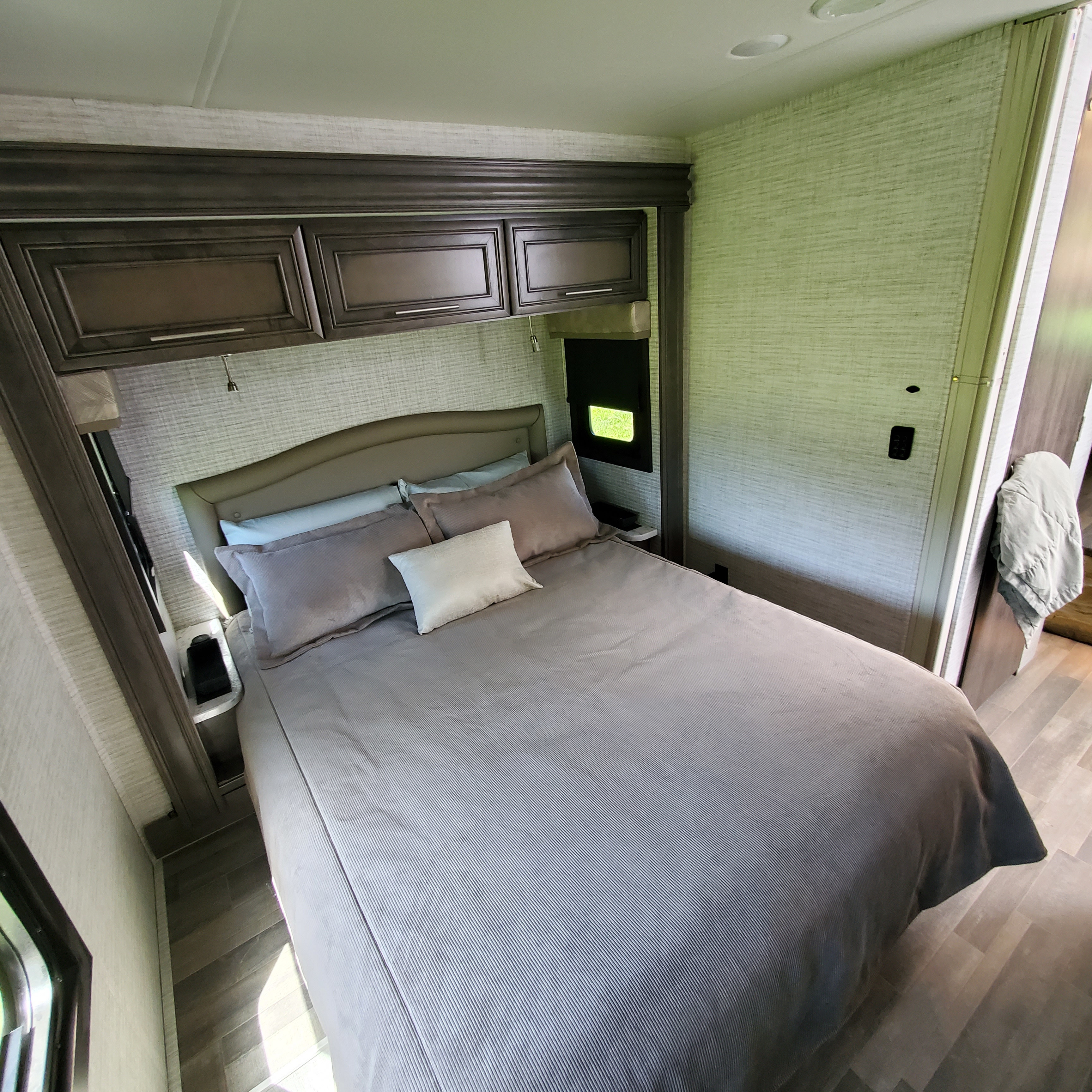 Cozy rv rental bedroom features a queen bed with grey bedding, overhead cabinets, and a window for comfortable travel.. Jayco Greyhawk 2024