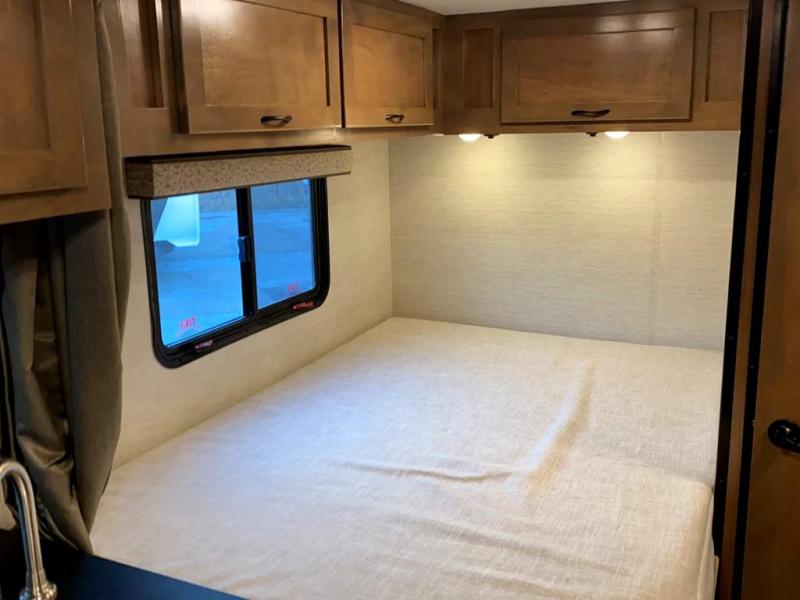 Inside an RV rental: A comfy bed with light sheets, overhead cabinets, and a window. Under-cabinet lights illuminate the wall.. Tiffin Motorhomes Wayfarer 2021