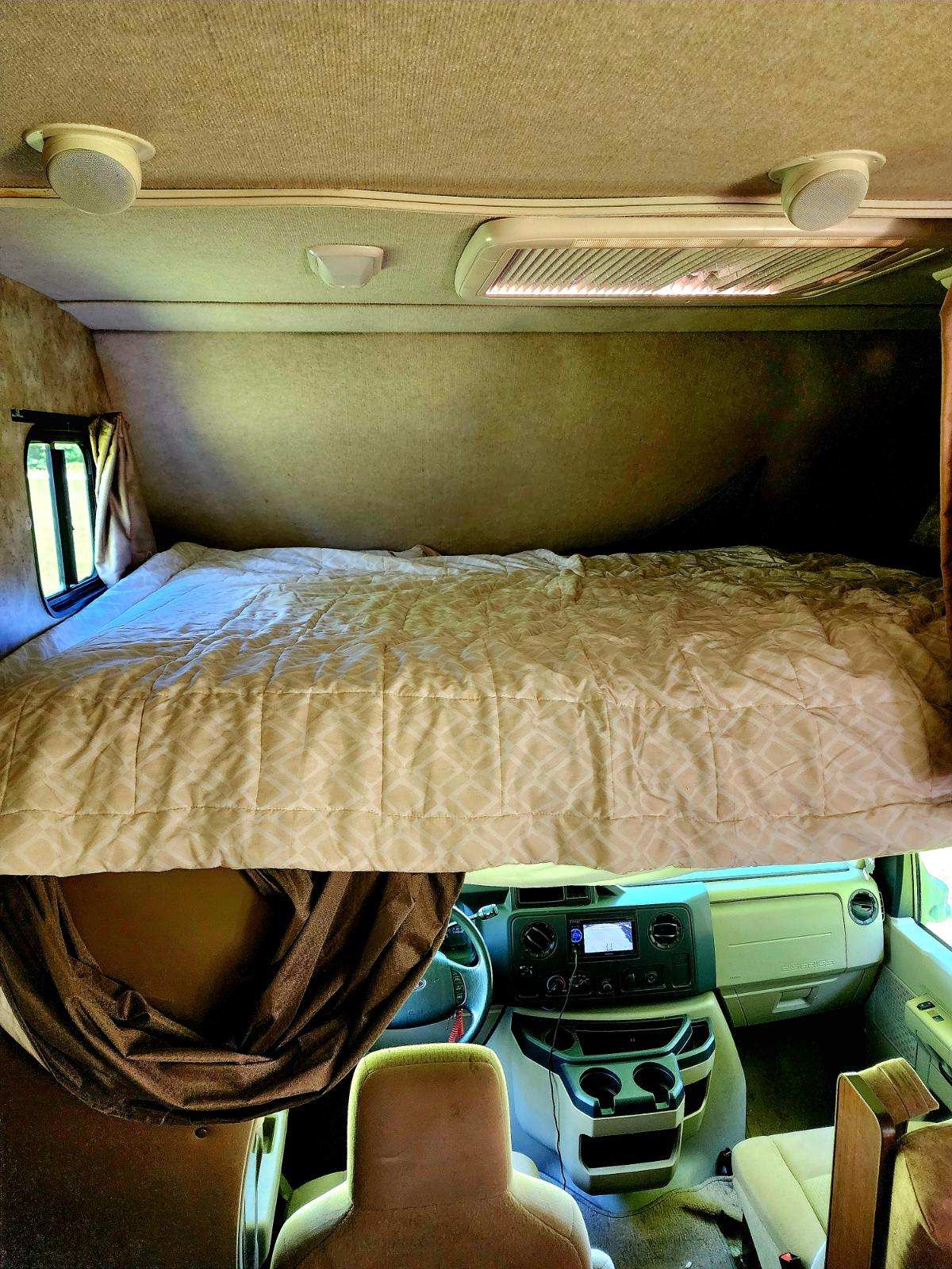 Cozy over-cab bed with a quilted comforter in this RV rental, overlooking the driver's cockpit and dashboard.. Tioga Ranger 2013