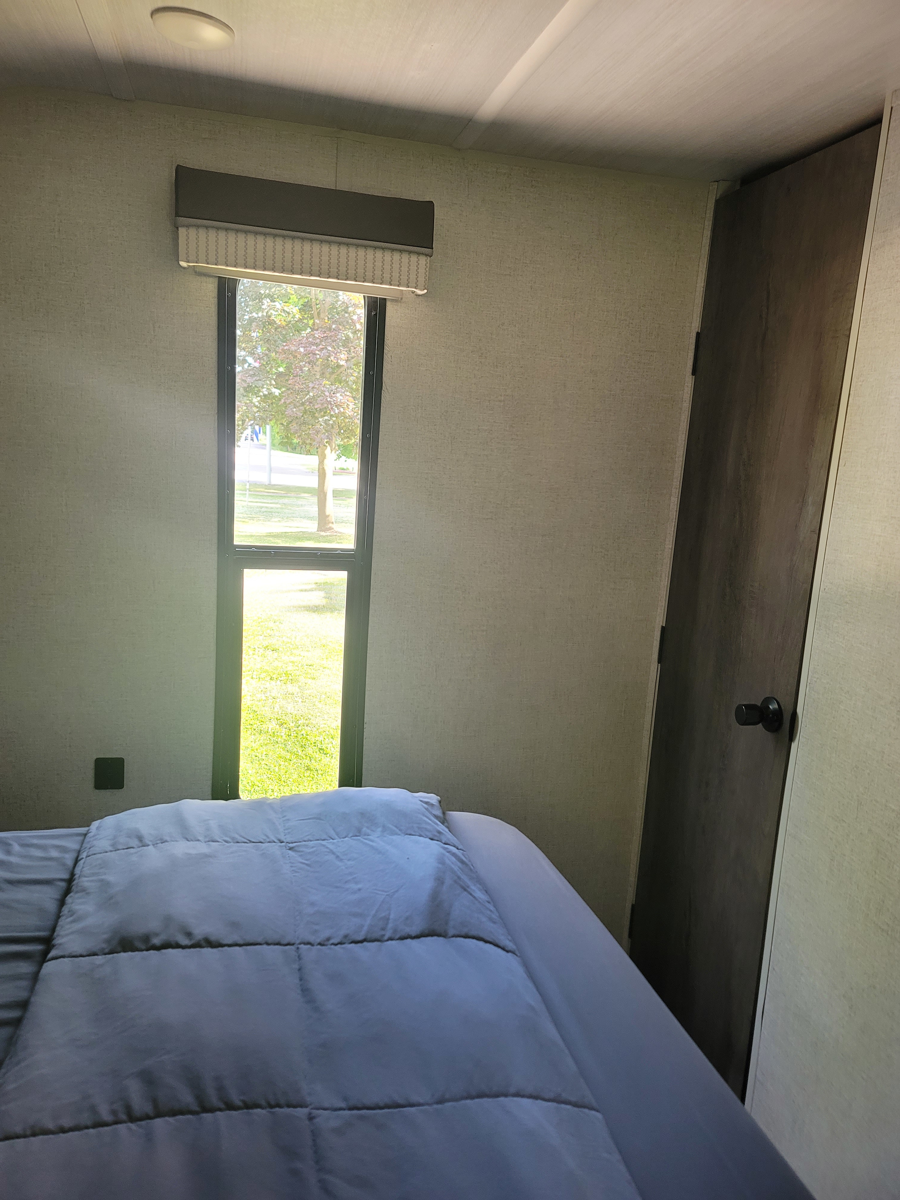 Inside an RV rental bedroom: A comfortable bed, a narrow window offering a vibrant green outdoor view, and a nearby door.. Forest River Salem 2021
