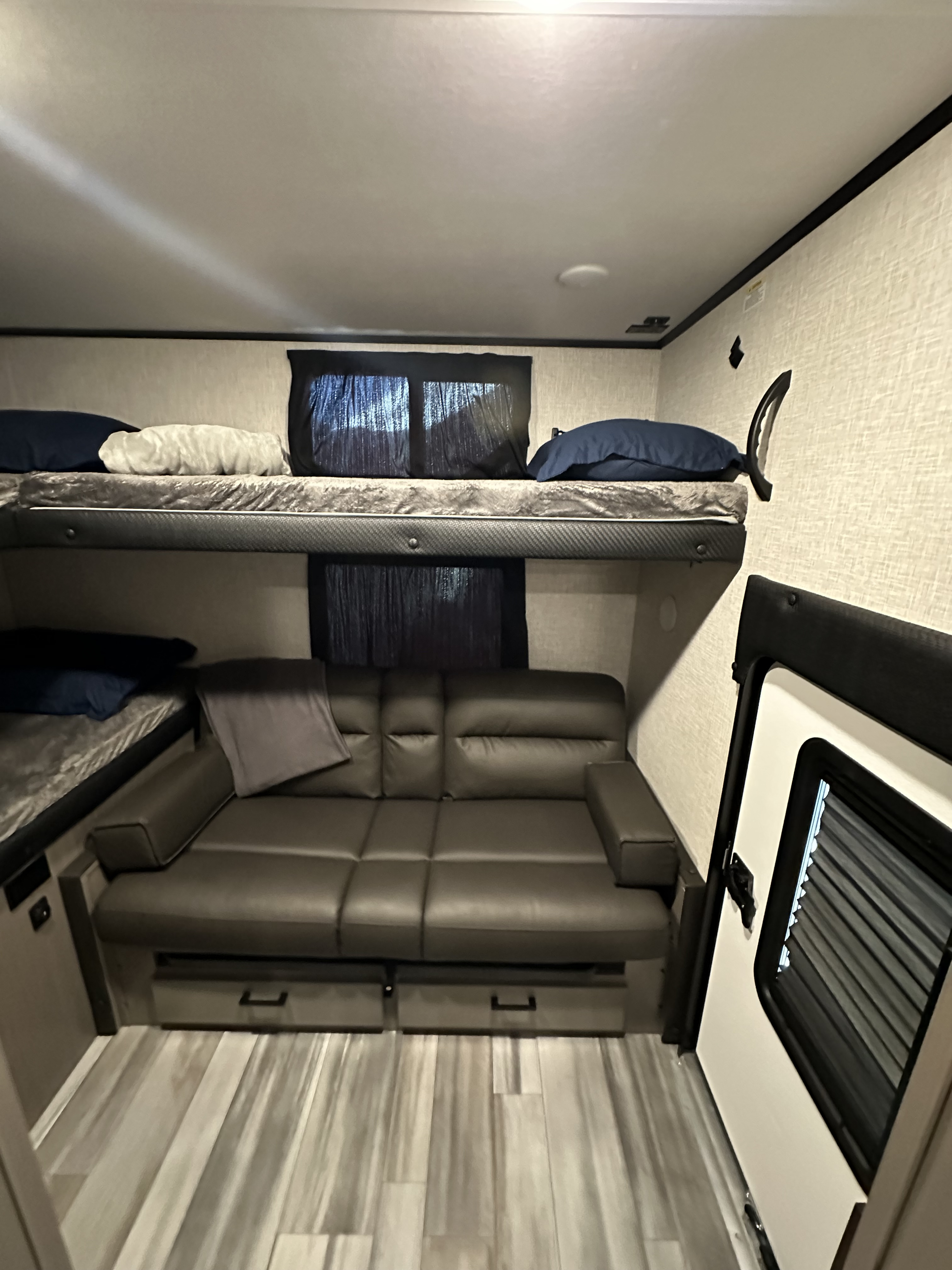 Spacious RV rental interior featuring cozy bunk beds with pillows, a grey sofa, and durable flooring. Entry door open.. Jayco Jay Feather 2024