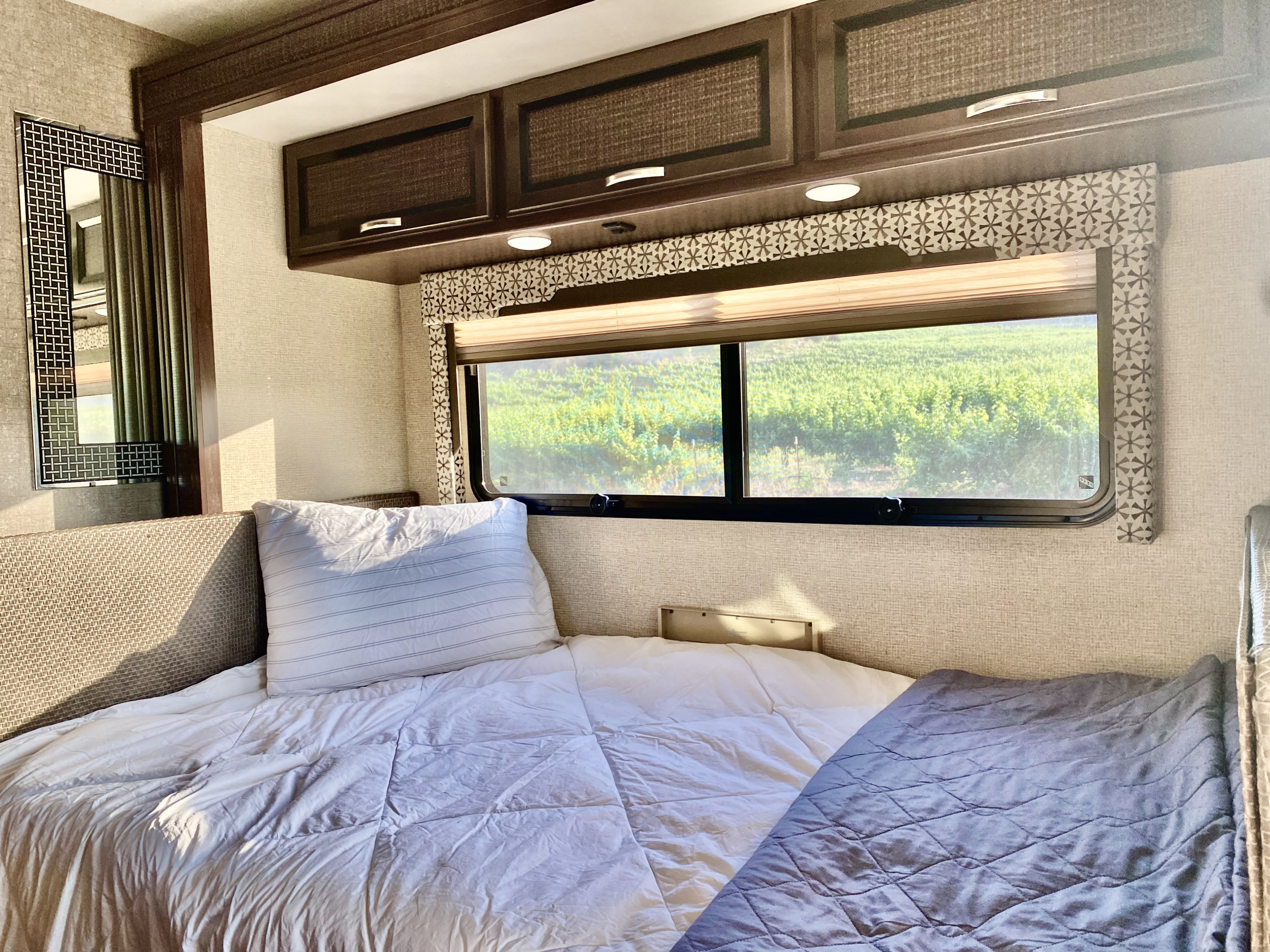 Cozy RV rental bedroom with a comfy bed, overhead cabinets, and a scenic window view of green vineyards.. Thor Axis 2017