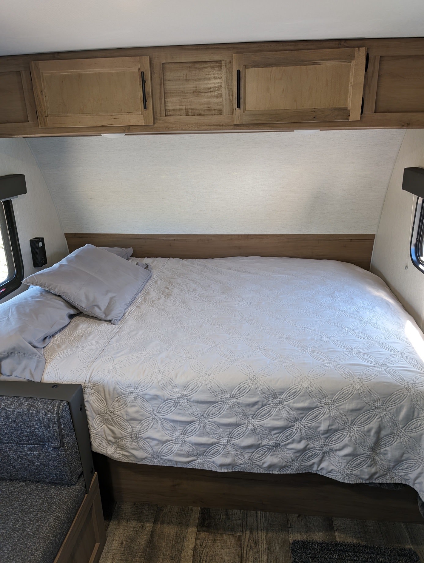 An RV rental's bedroom shows a neat bed with light bedding, gray pillows, and wooden overhead storage cabinets.. Forest River Cherokee Wolf Pup 2023