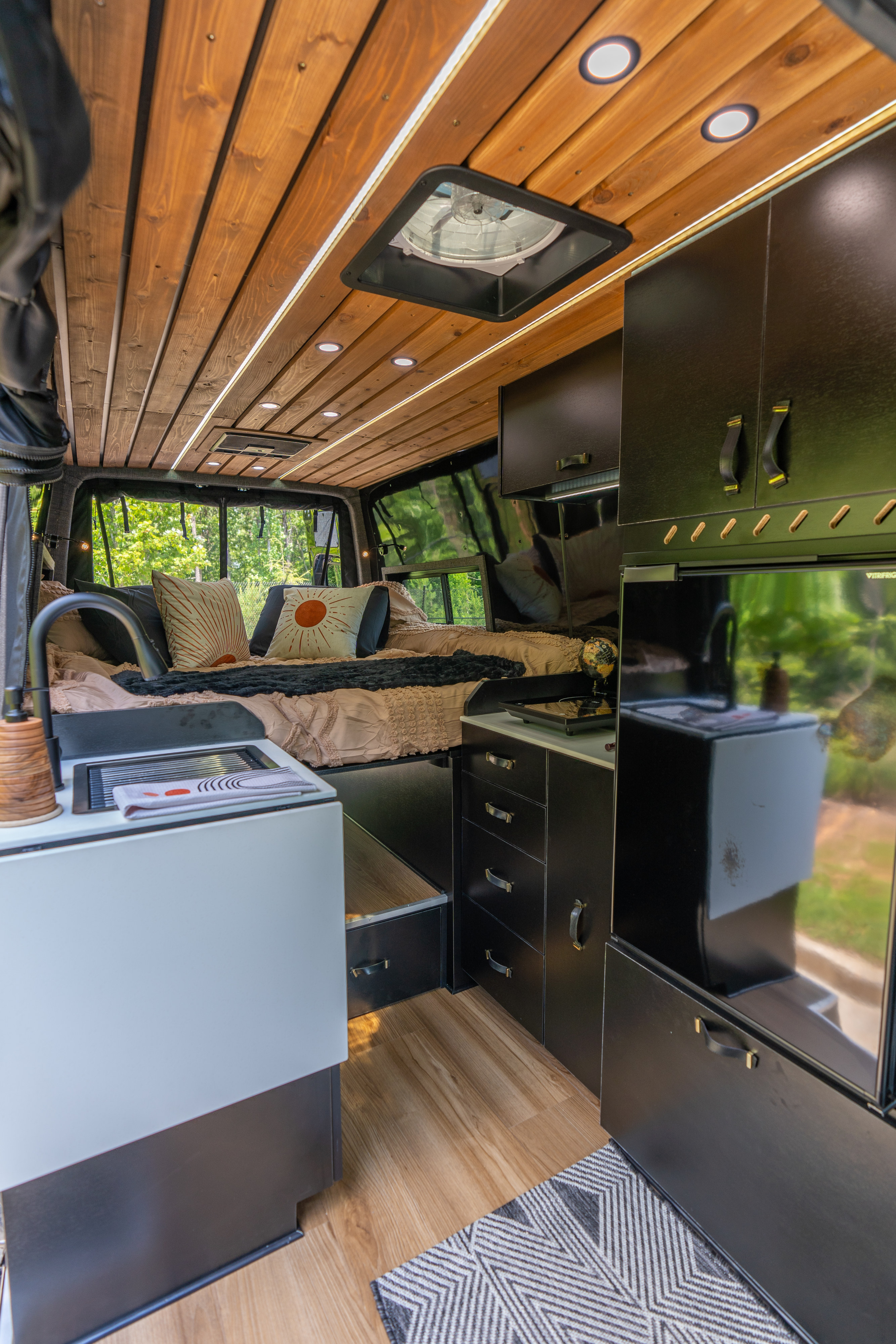 Explore this stylish RV rental's cozy interior, featuring a wooden ceiling, built-in black cabinets, and a compact kitchen.. Mercedes-Benz Sprinter 2023