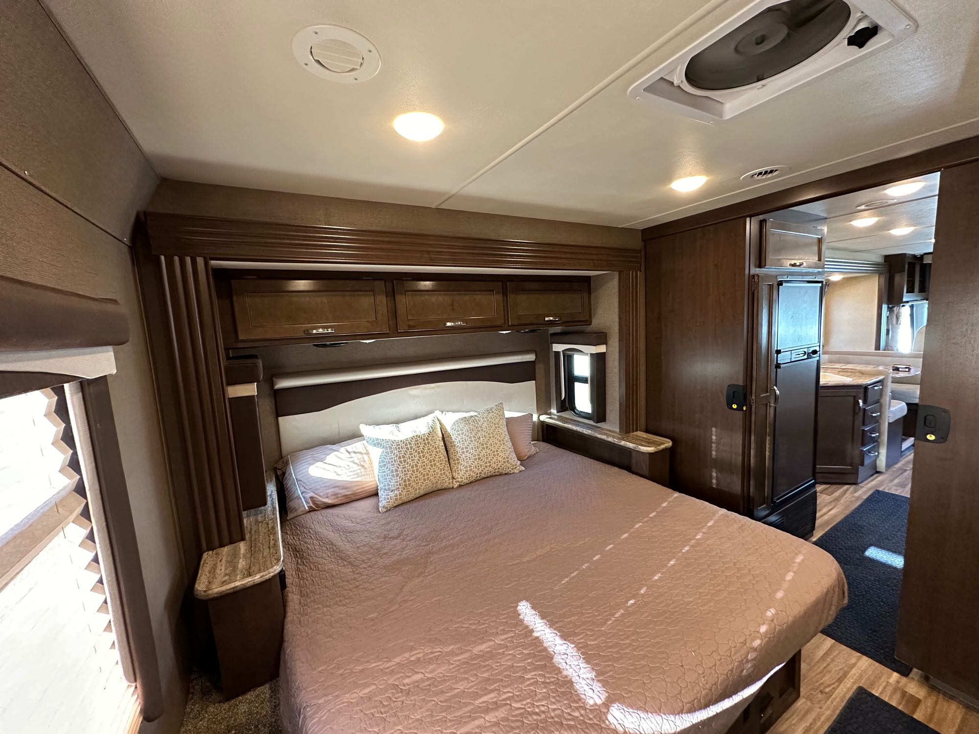 Spacious RV rental bedroom: large bed, dark wood cabinetry, and a view into the modern kitchen area.. Thor A.C.E. 2017