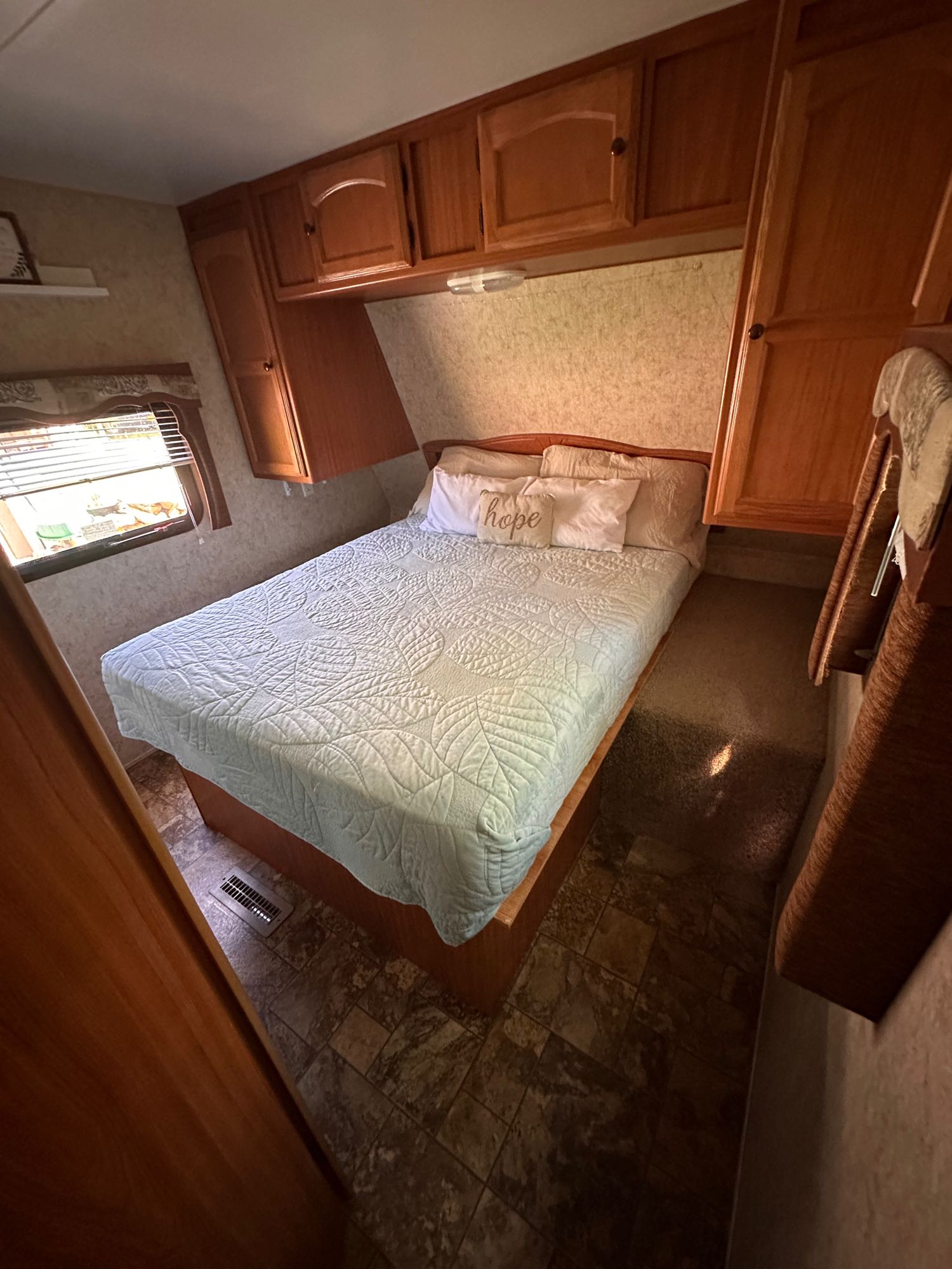 RV rental bedroom: Bed with light blue quilt, overhead wooden cabinets, and window with blinds. Cozy interior.. Four Winds Breeze SS 2010