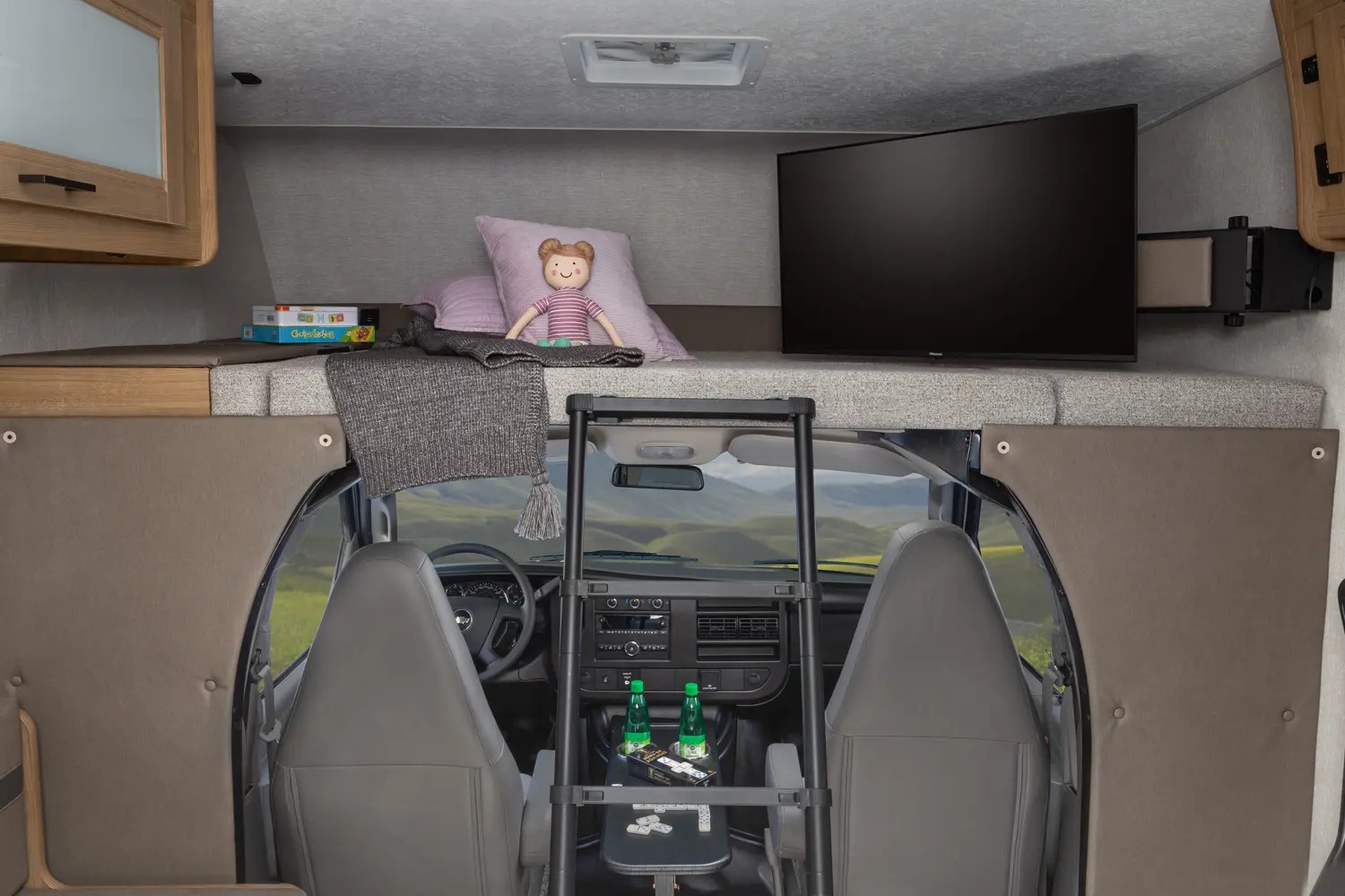 Spacious RV rental interior featuring a cozy overhead bunk with bedding and games, a TV, and a view into the front cab.. Forest River Coachmen Leprechaun 2024