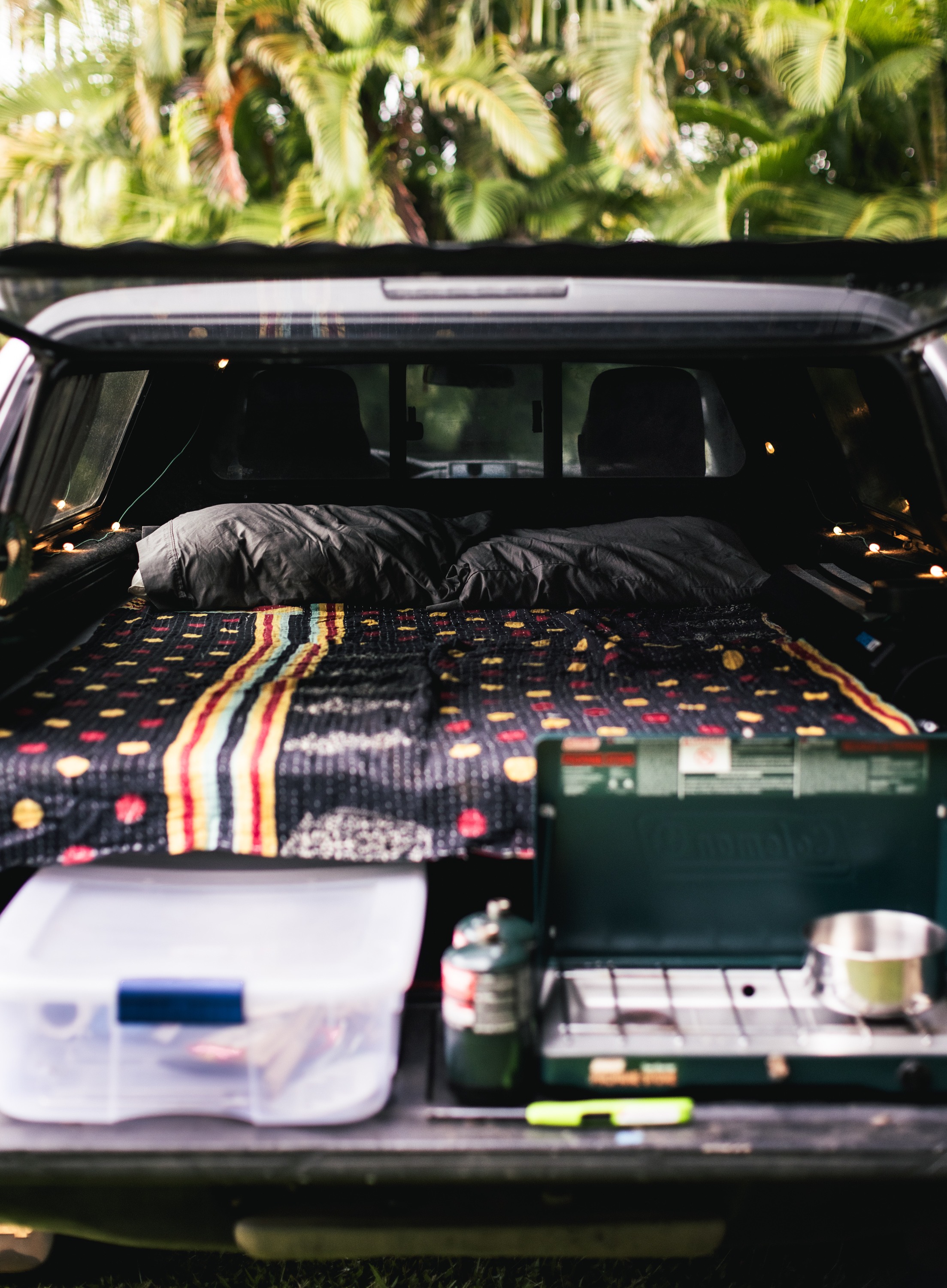 Cozy RV rental truck bed setup with colorful blanket, pillows, fairy lights, and a camp kitchen for outdoor adventure.. toyota tacoma 2012