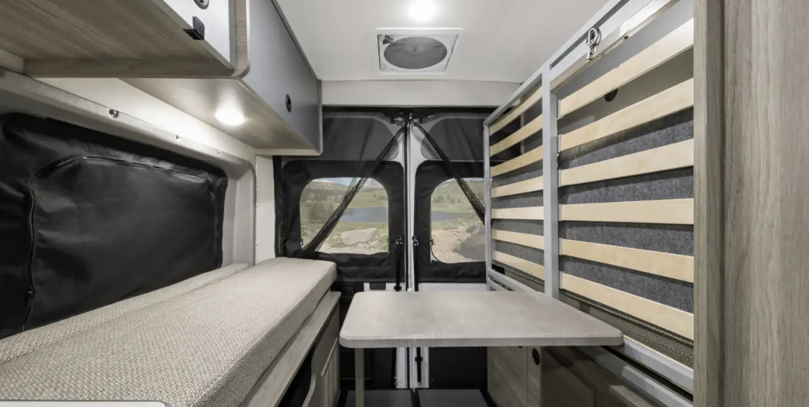 This RV rental interior features a convertible bed, dining table, and screened rear doors showcasing a beautiful lake view.. Winnebago Solis 2021