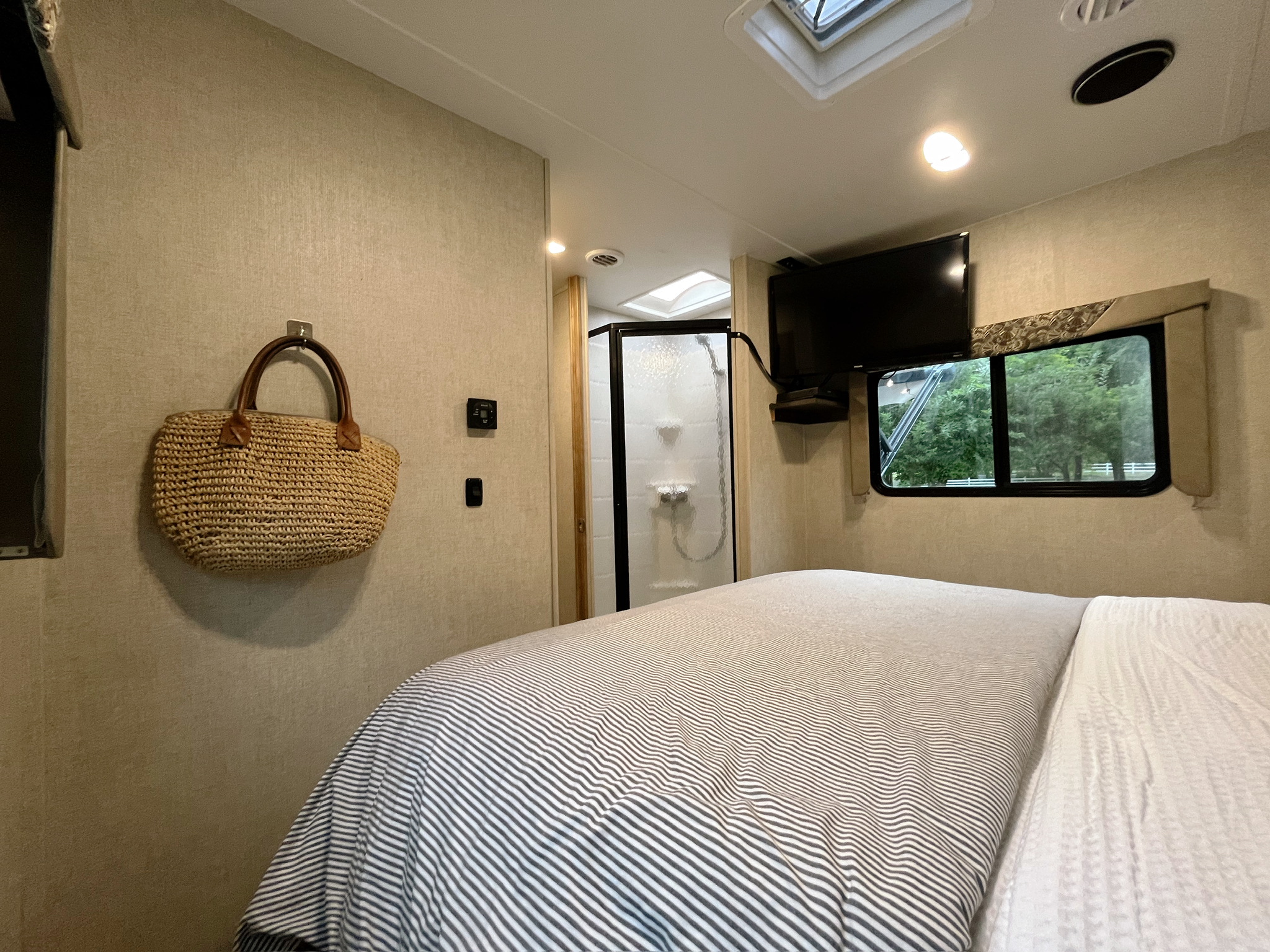 Cozy RV rental bedroom featuring a striped bed, wall-mounted TV, window view of nature, and an ensuite shower.. Gulf Stream Conquest 2017