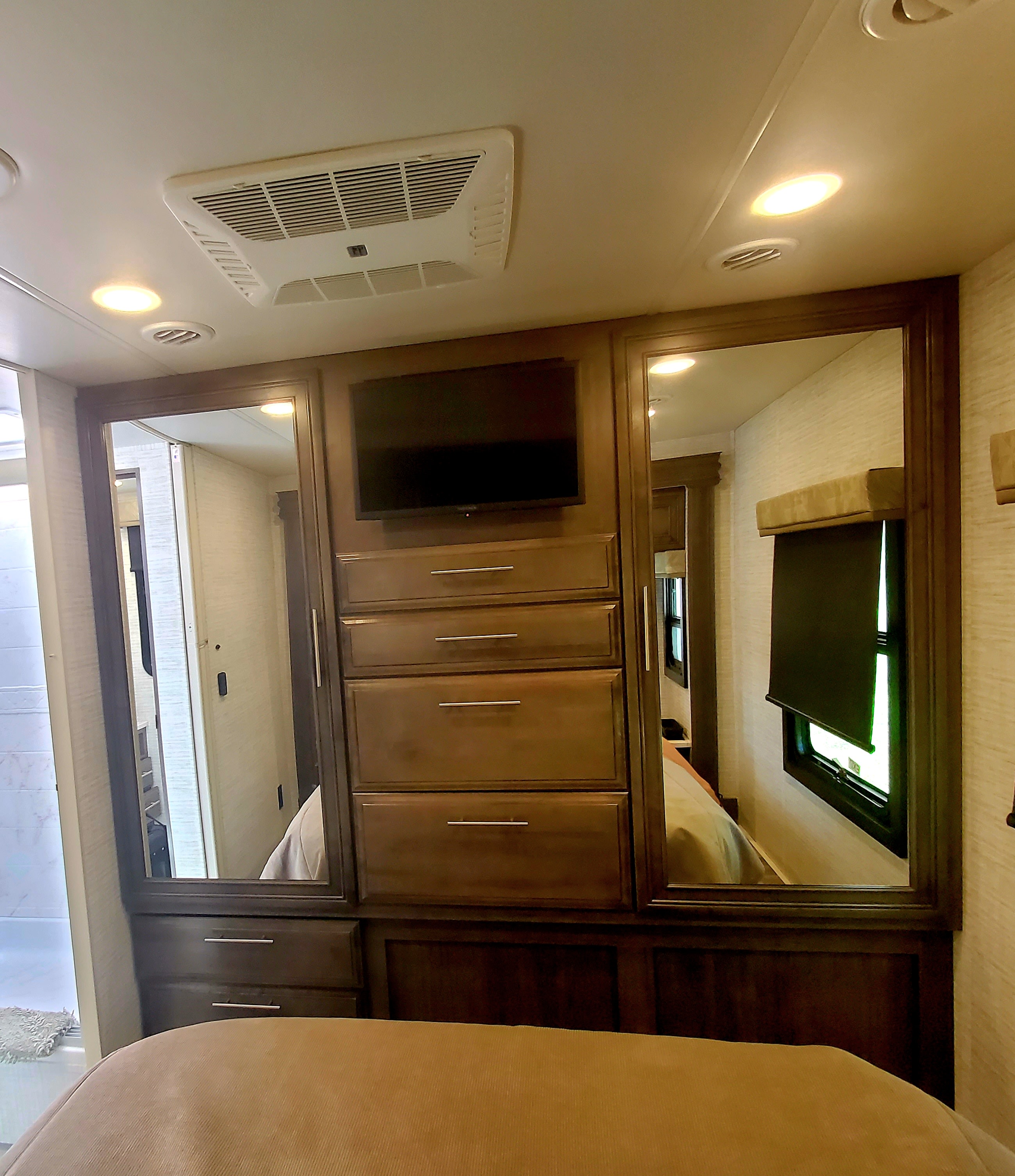 This RV rental bedroom features mirrored wardrobe doors flanking a dresser with a TV. Overhead AC and lighting complete the compact space.. Jayco Greyhawk 2024