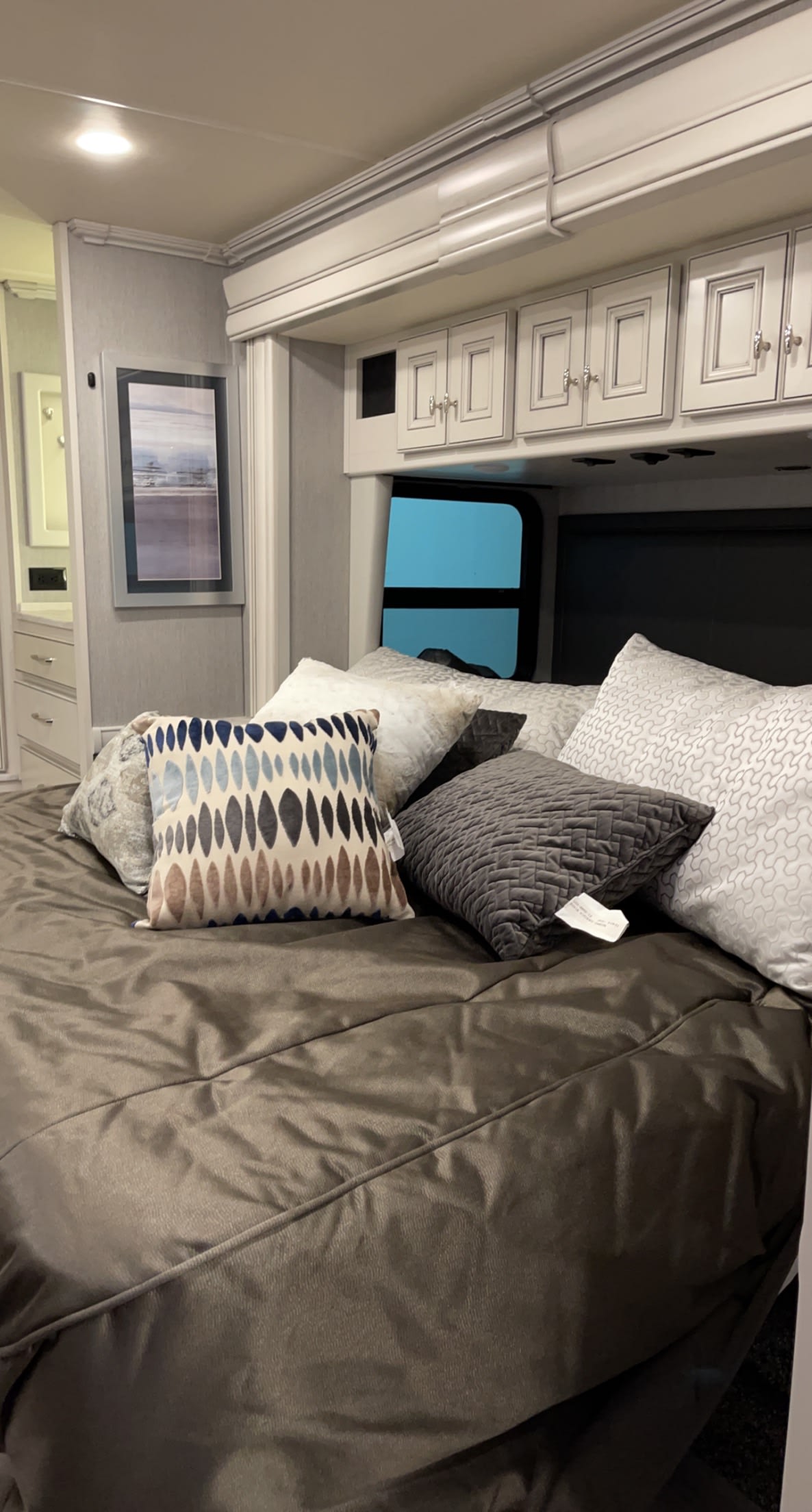 A modern RV rental bedroom featuring a made bed with multiple pillows, overhead cabinets, and a framed picture.. Holiday Rambler Nautica 2022