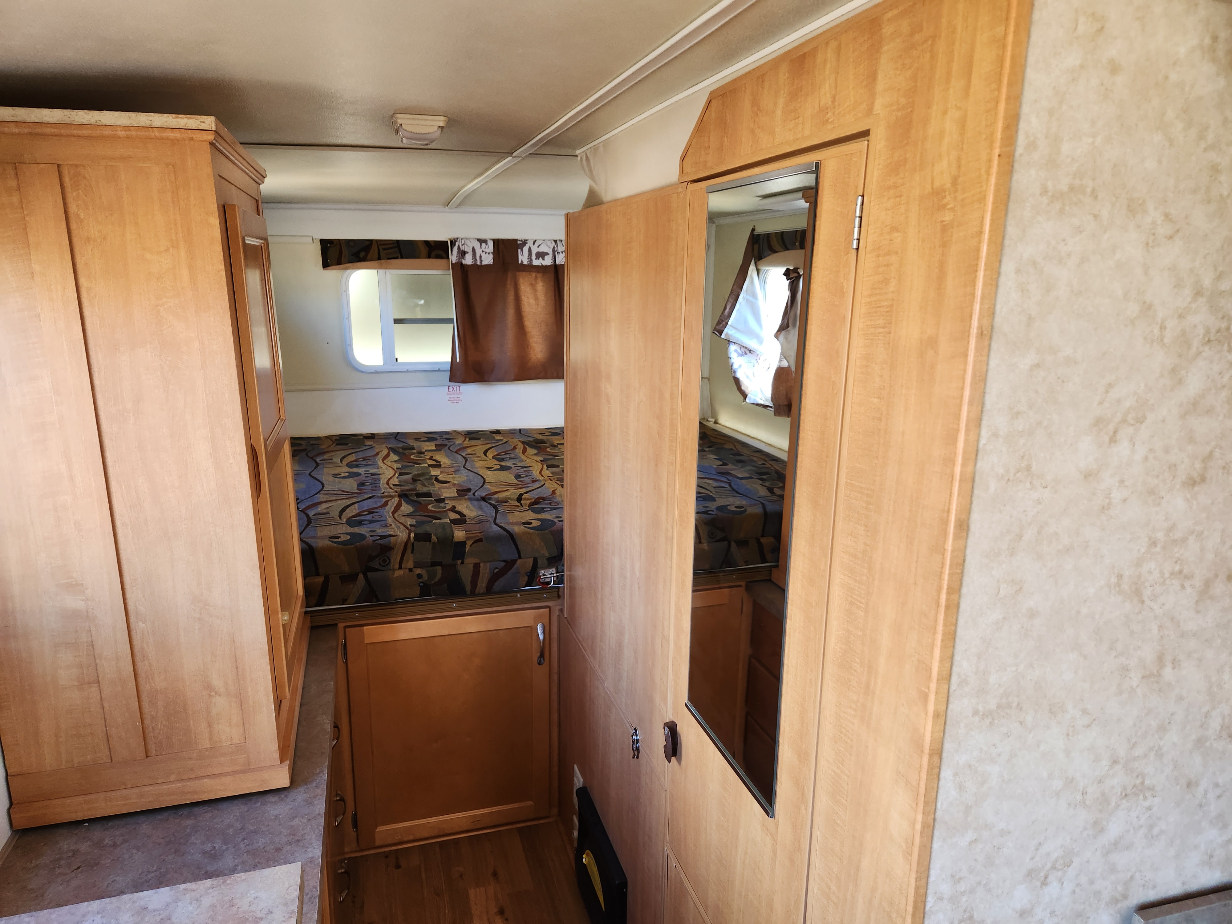 Compact RV rental bedroom showcasing functional wood cabinetry, a bed with patterned cover, window, and mirrored wardrobe.. Trailmanor Trailmanor Trailer 2009