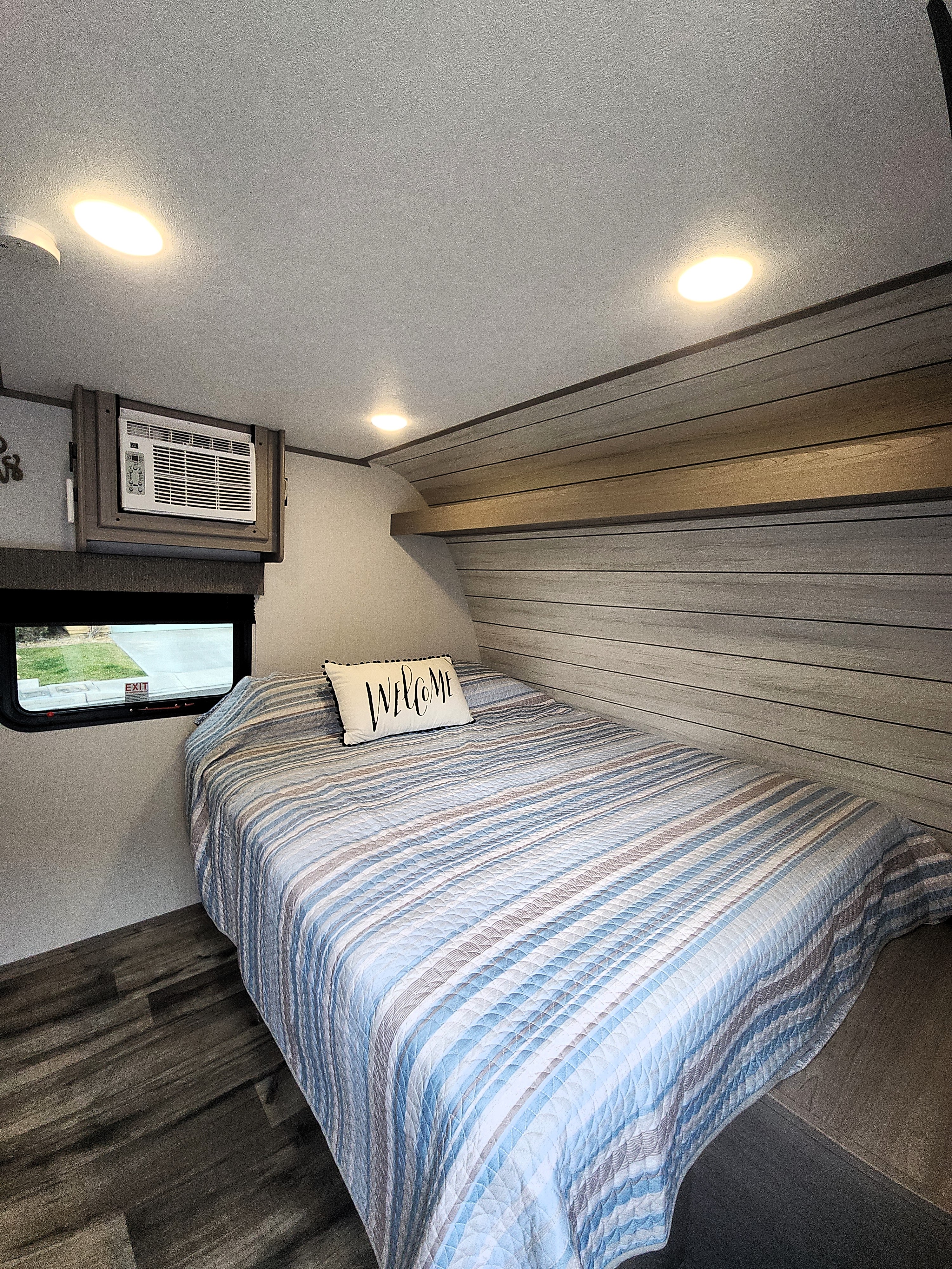 Cozy RV rental bedroom with striped bed, "Welcome" pillow, window, AC unit, and wood-paneled walls.. Colemen Dutchmen 17R 2024