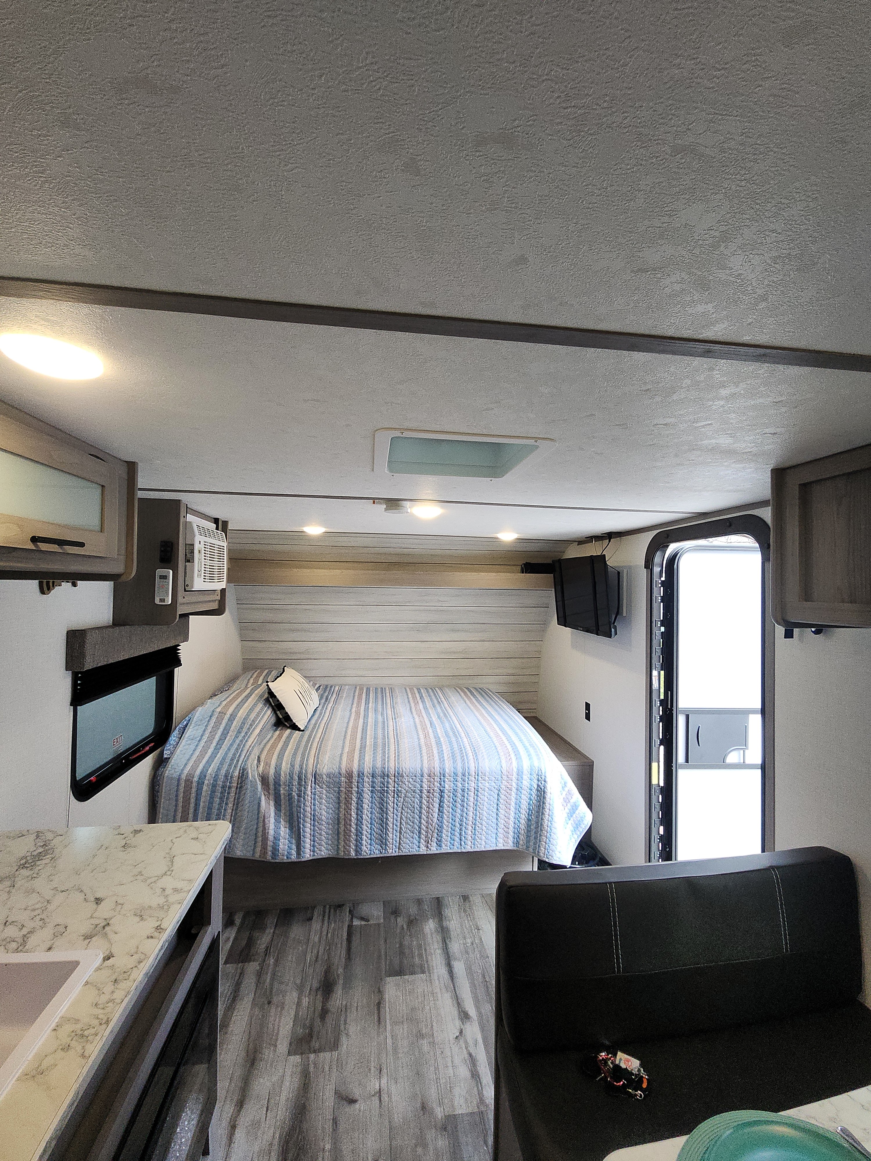 RV rental interior showcasing a cozy bedroom with a striped quilt, compact kitchen, and modern wood-look flooring.. Colemen Dutchmen 17R 2024