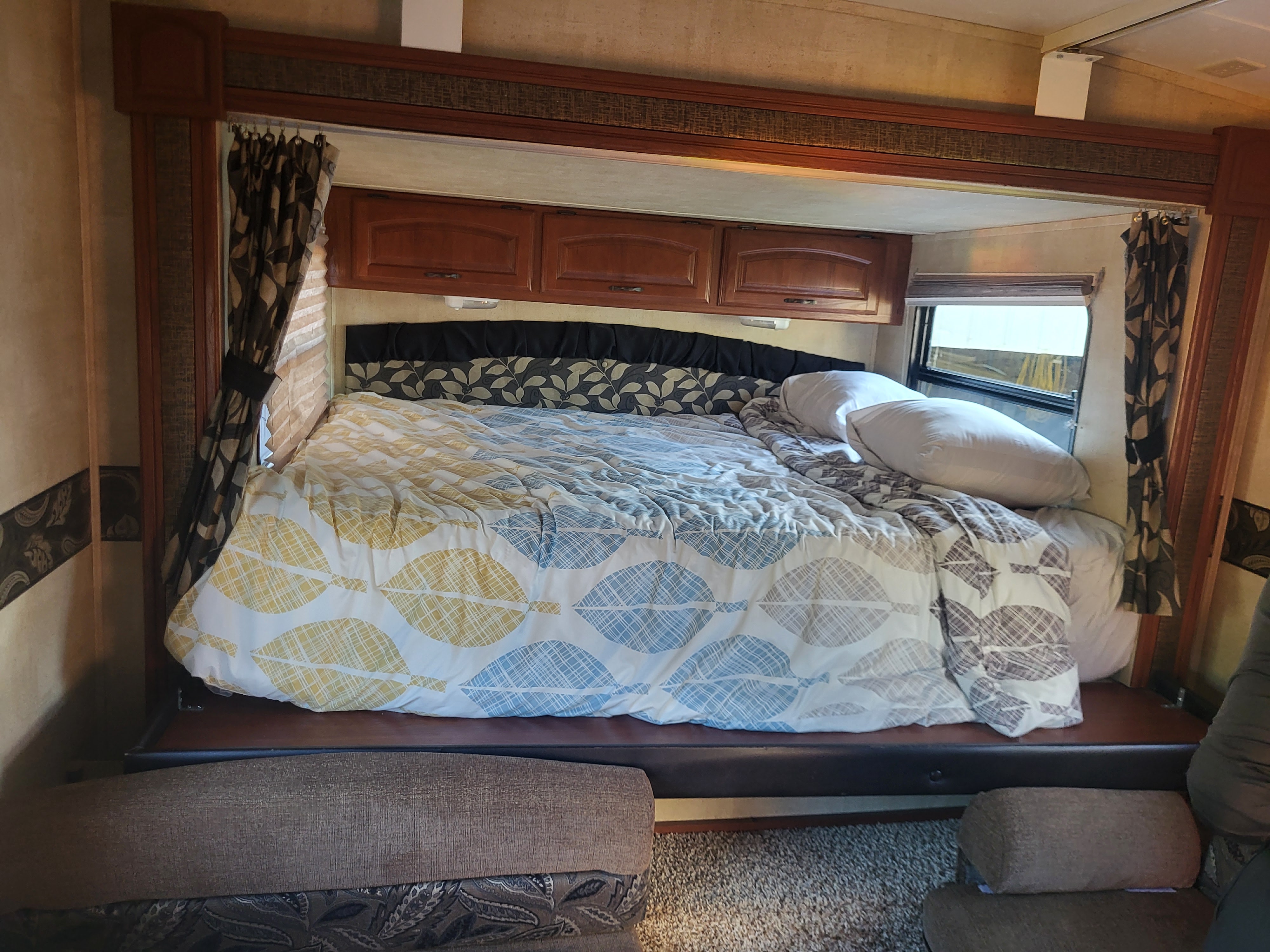 Inside an RV rental: a cozy bedroom setup featuring a leaf-patterned duvet, pillows, overhead cabinets, and window with curtains.. Keystone RV Outback 10th Anniversary 2012