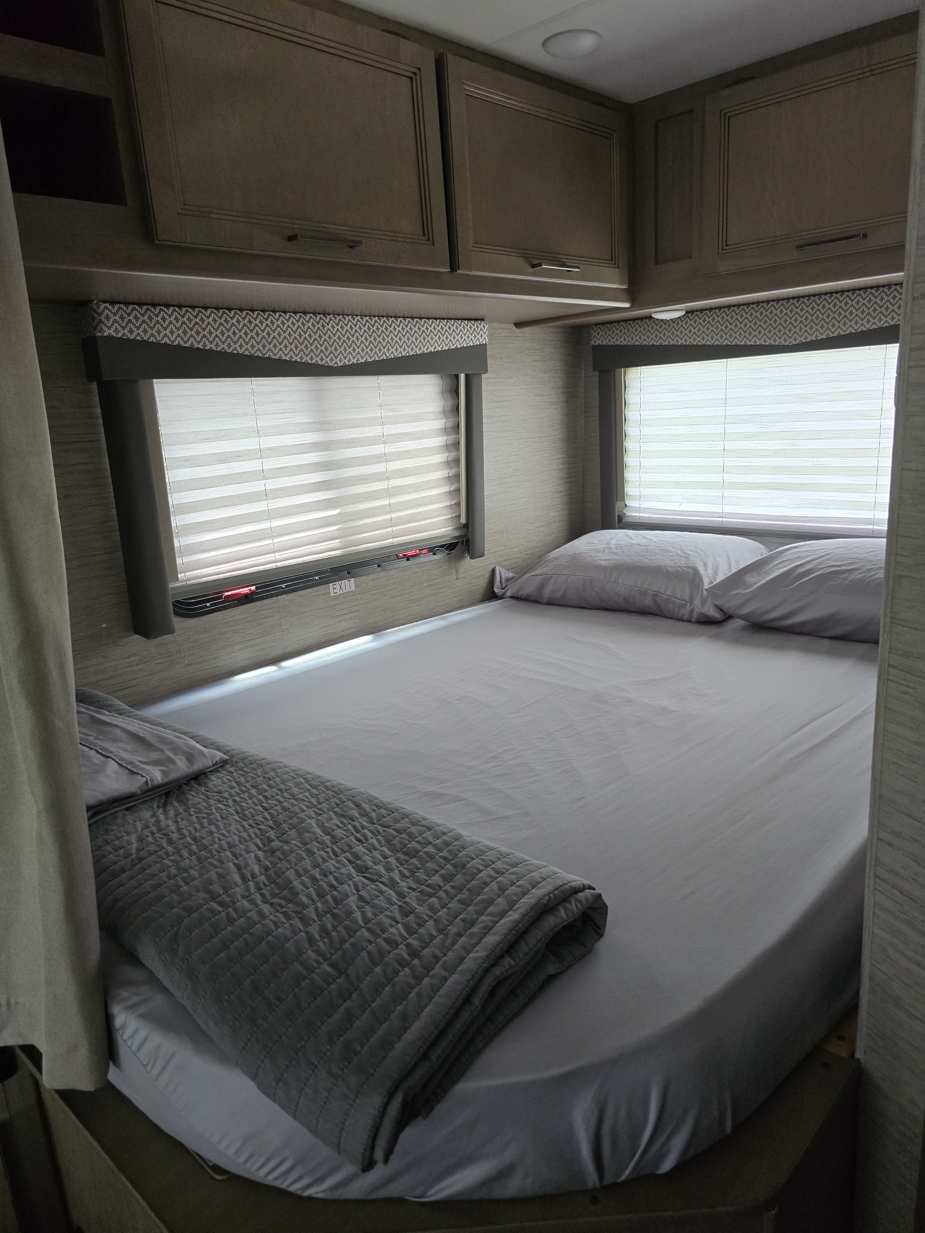 Inviting RV rental bedroom with a neatly made bed, light grey sheets, overhead cabinets, and bright windows.. Thor Four Winds 2022