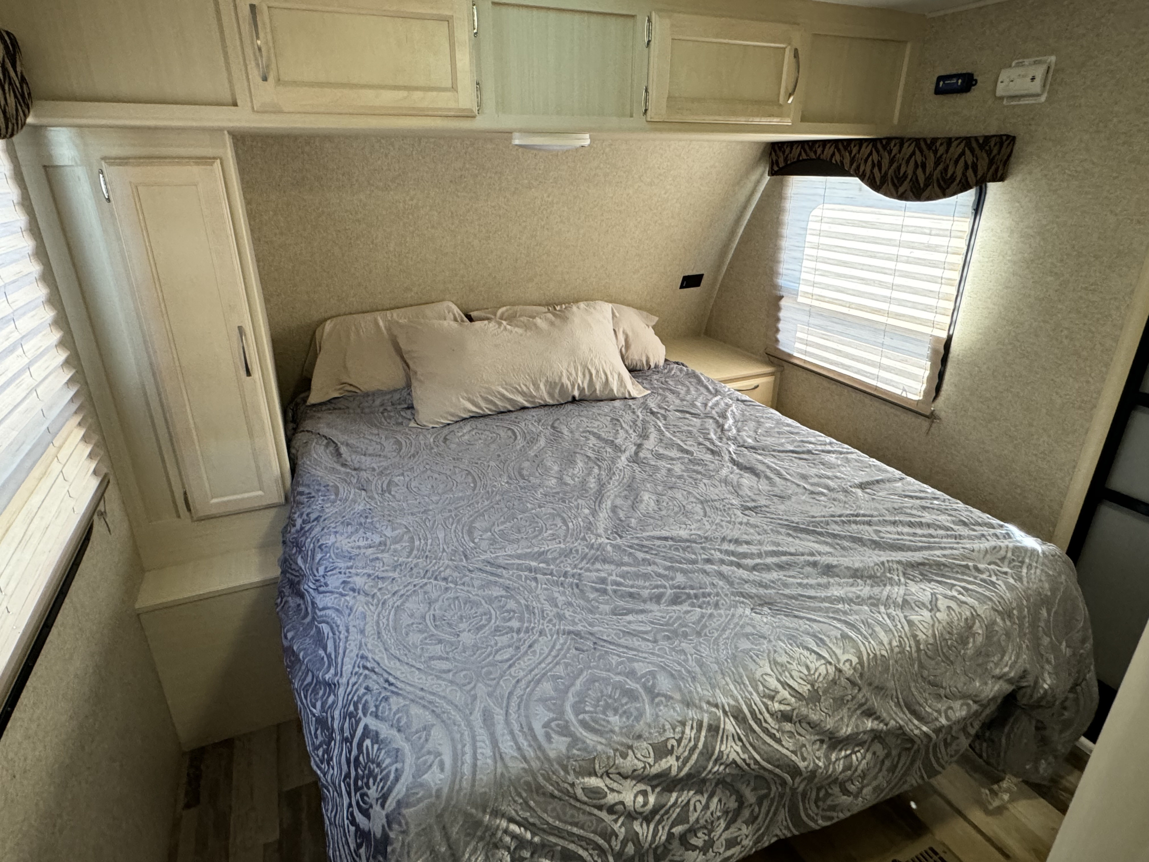 Cozy RV rental bedroom featuring a large bed with a patterned comforter, ample overhead cabinets, and window with blinds.. Winnebago Minnie Winnie 2017