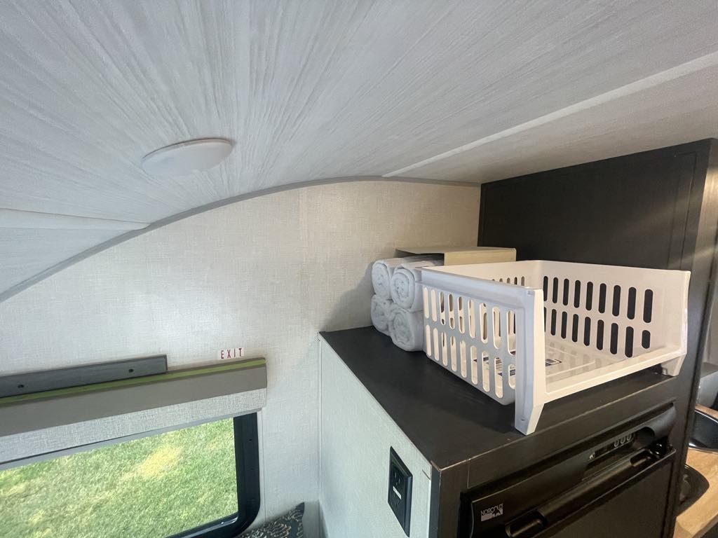 Inside an RV rental, showing compact living space: ceiling, window, "EXIT" sign, storage basket, towels, and a mini-fridge below.. R-Pod R-Pod Trailer 2023