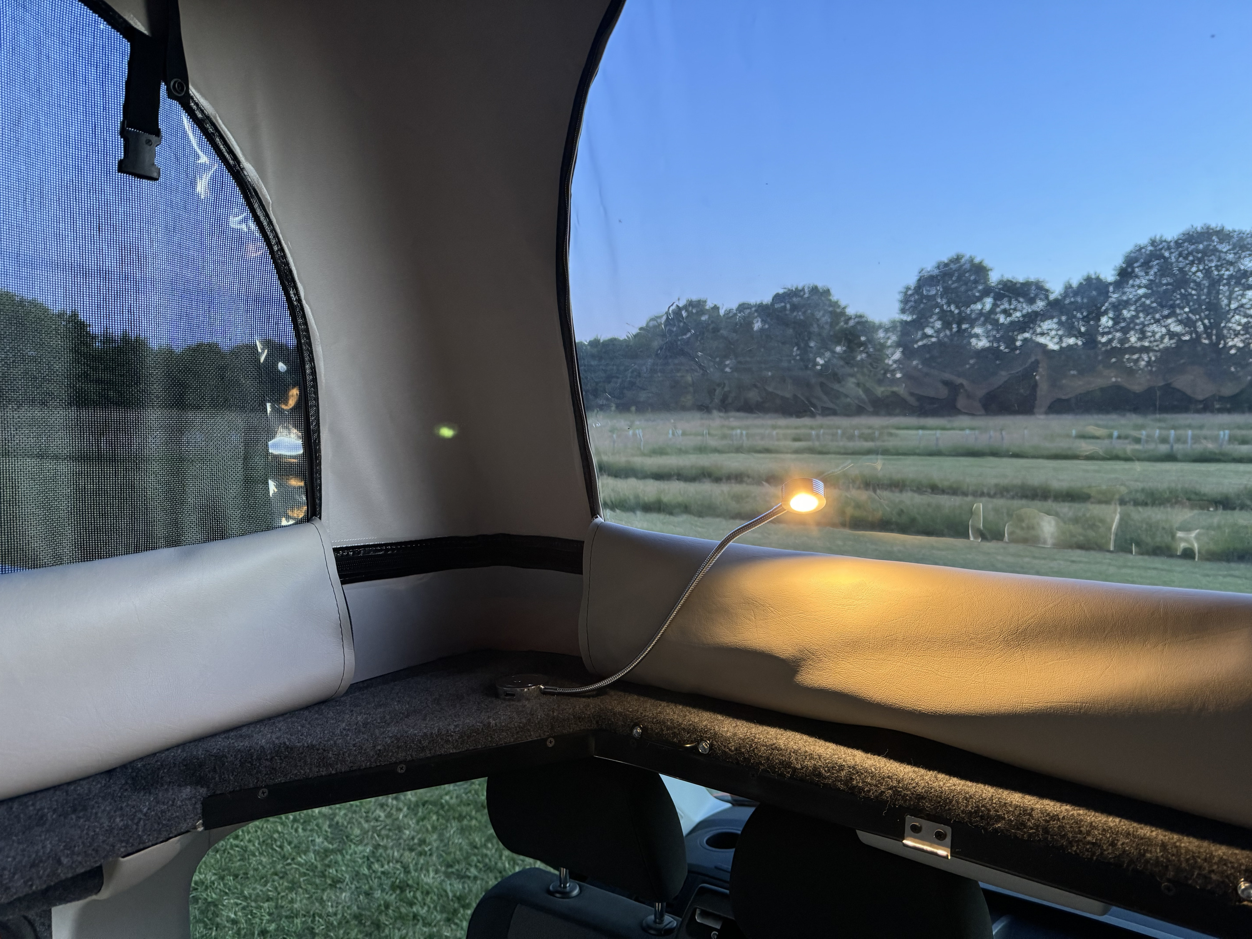 Interior view of an rv rental pop-top, showing mesh windows, a bright reading light, and an exterior green field.. Volkswagen T6 transporter 2018