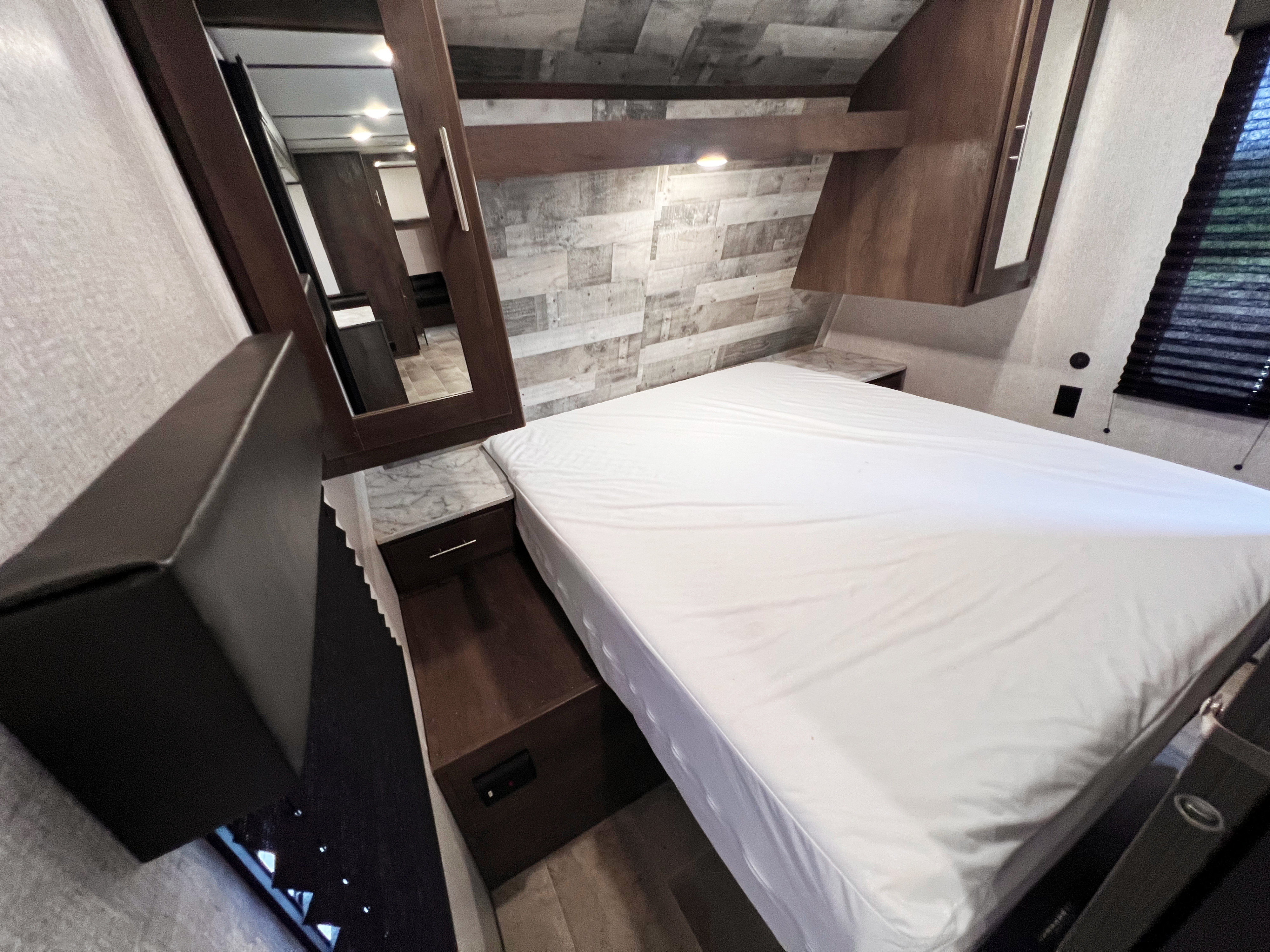 RV rental bedroom view: a white bed, wood-paneled wall, mirrored cabinet, and bedside table.. Dutchmen Lantern 2021