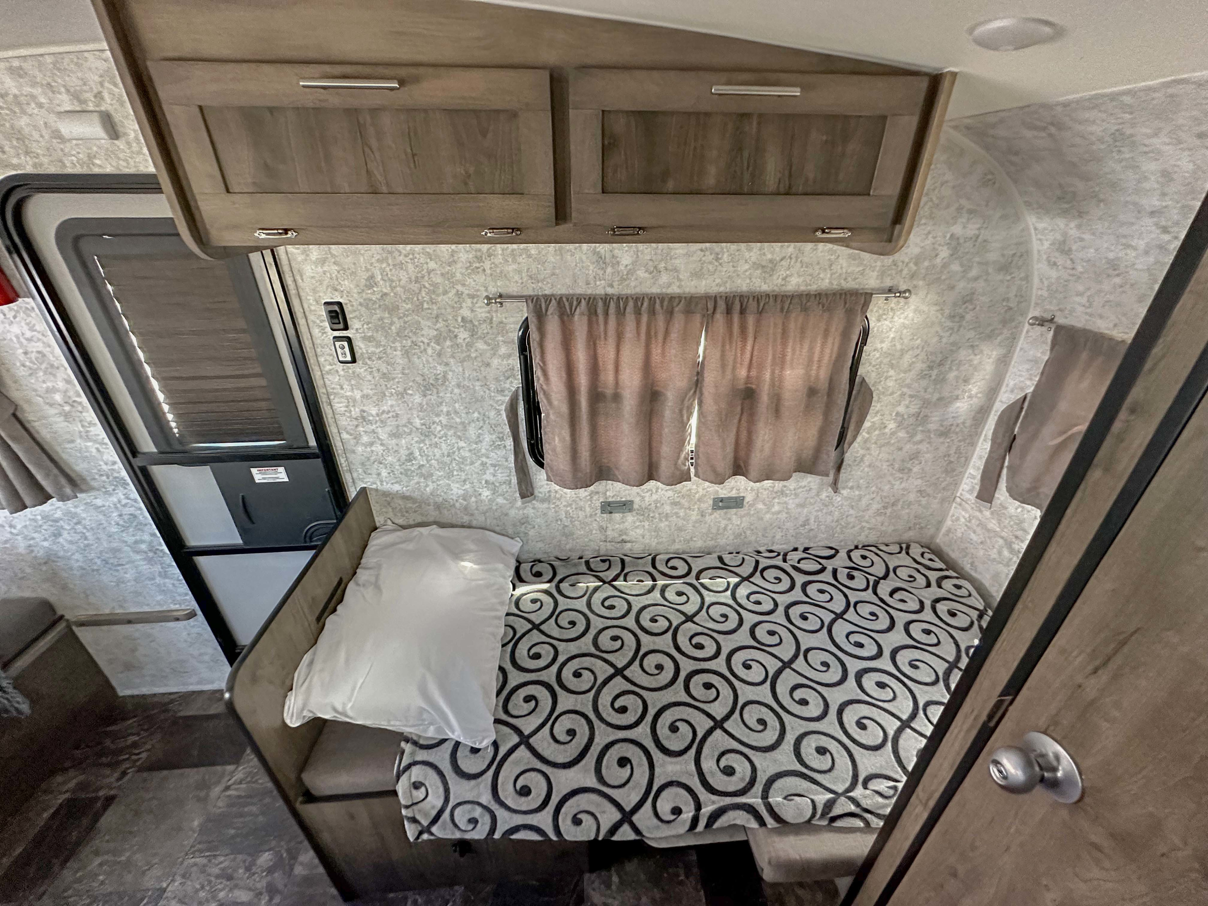 Explore a cozy RV rental interior, featuring a compact bedroom with a patterned bed, overhead cabinets, and window with curtains.. Prolite Lounge 2024