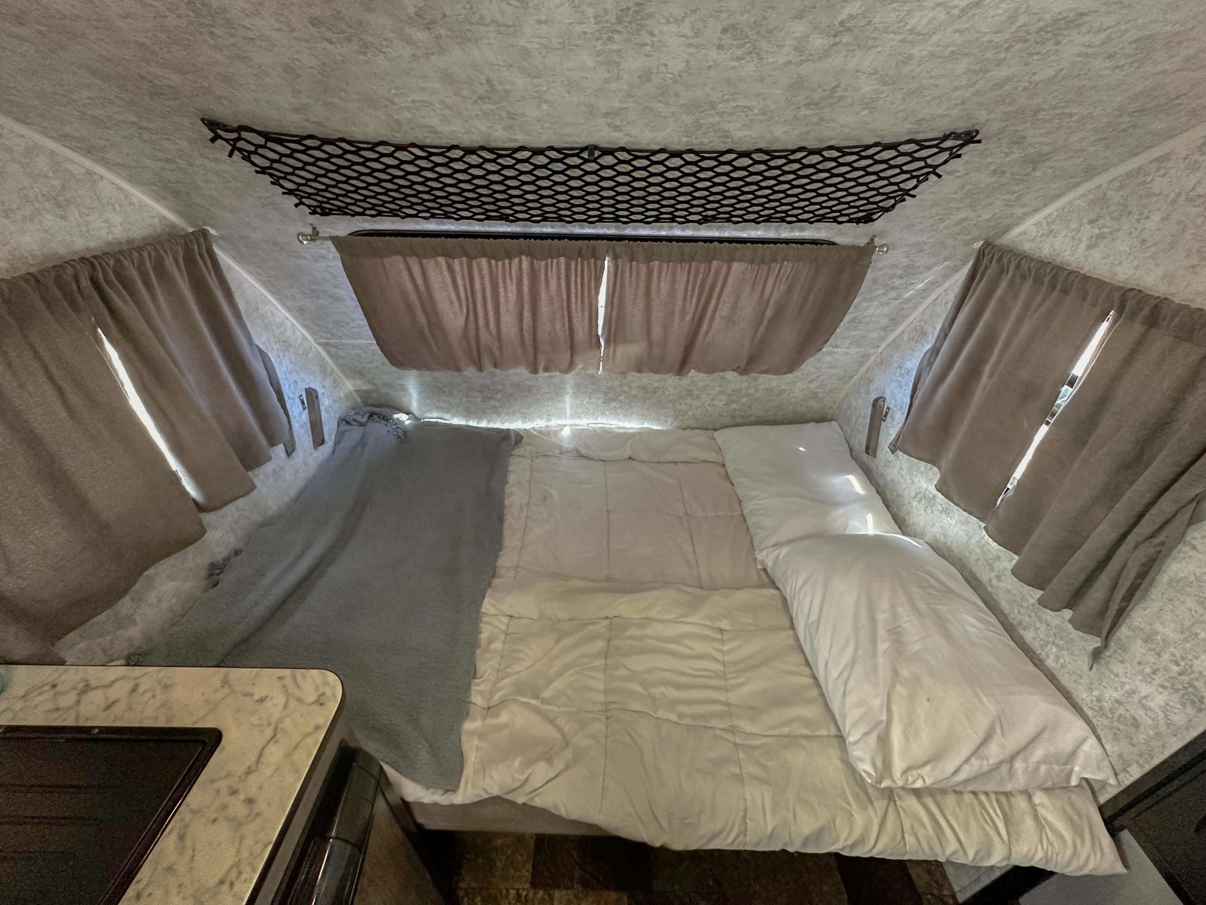 Inside this RV rental: a compact sleeping area with white bedding, a gray blanket, side curtains, and an overhead storage net.. Prolite Lounge 2024
