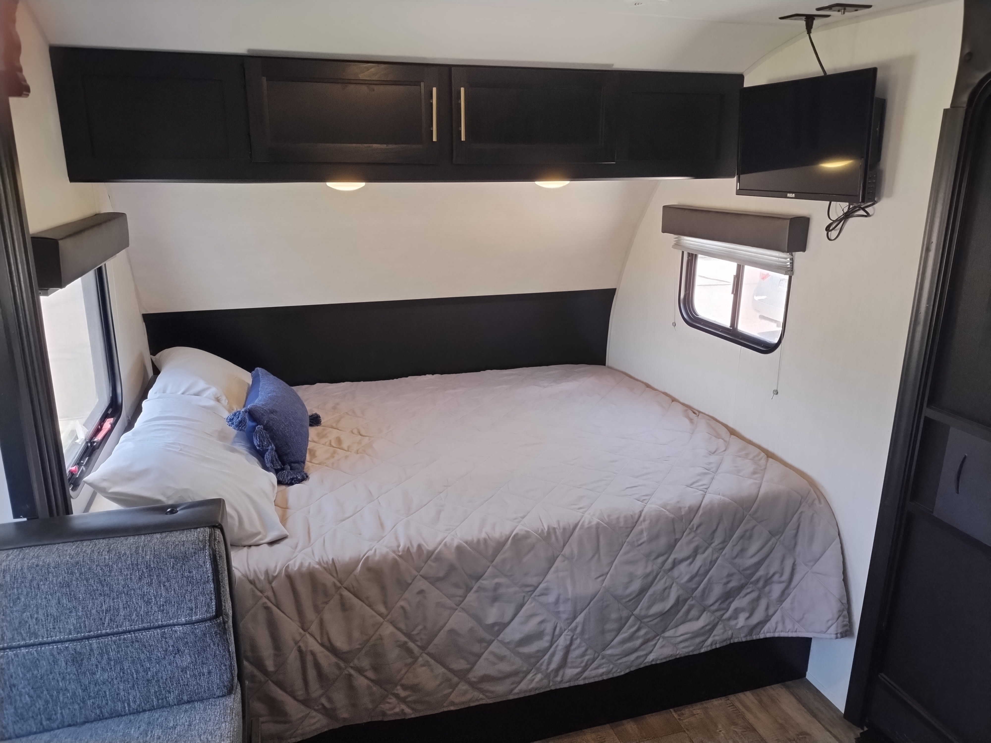 RV rental interior: a bedroom with a quilted bed, white pillows, dark overhead cabinets, window, and wall-mounted TV.. Forest River Cherokee Wolf Pup 2022