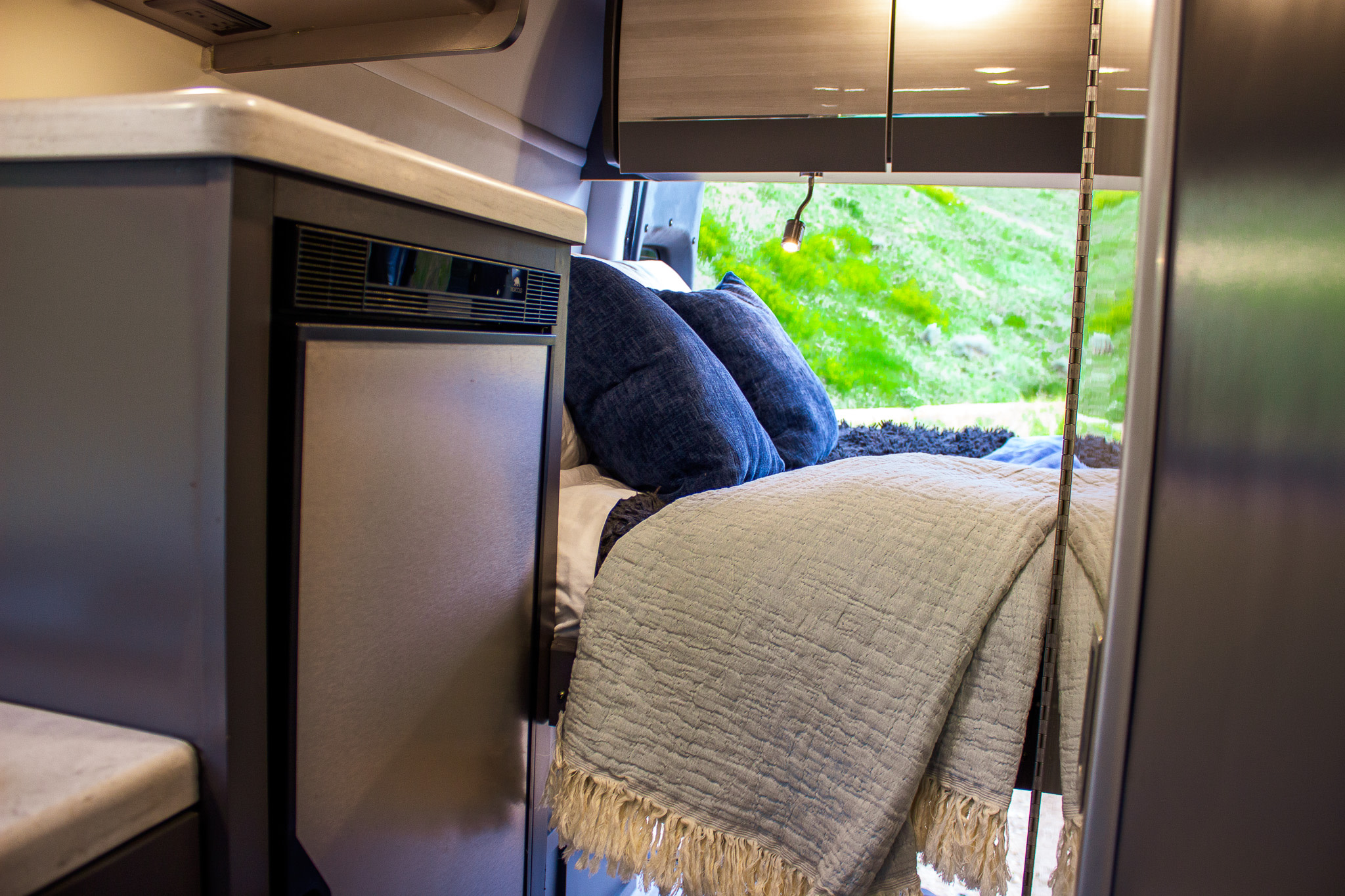 Compact RV rental interior features bed with blue pillows, a fridge, and scenic green views outside.. Mercedes-Benz Sprinter 4x4 2022