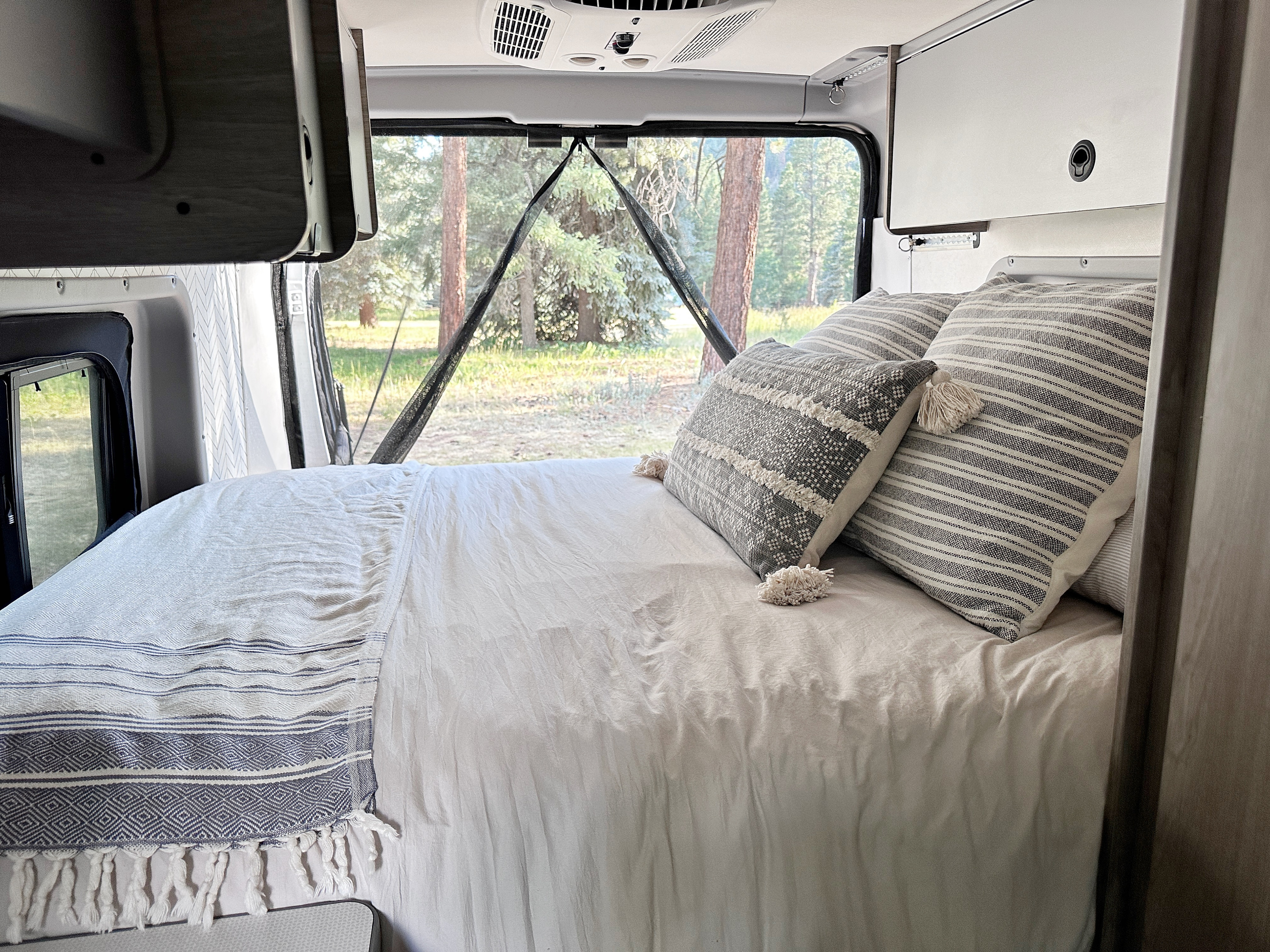Comfortable RV rental bed with decorative pillows, offering a peaceful forest view through open rear doors. Perfect for glamping.. Winnebago Solis 2022