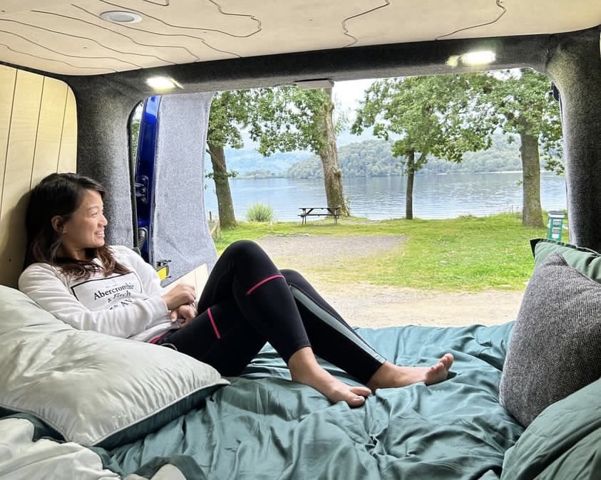 Serene lake view from a cozy RV rental. A woman relaxes on a bed inside a camper van, enjoying nature.. Ford Transit Custom 2016
