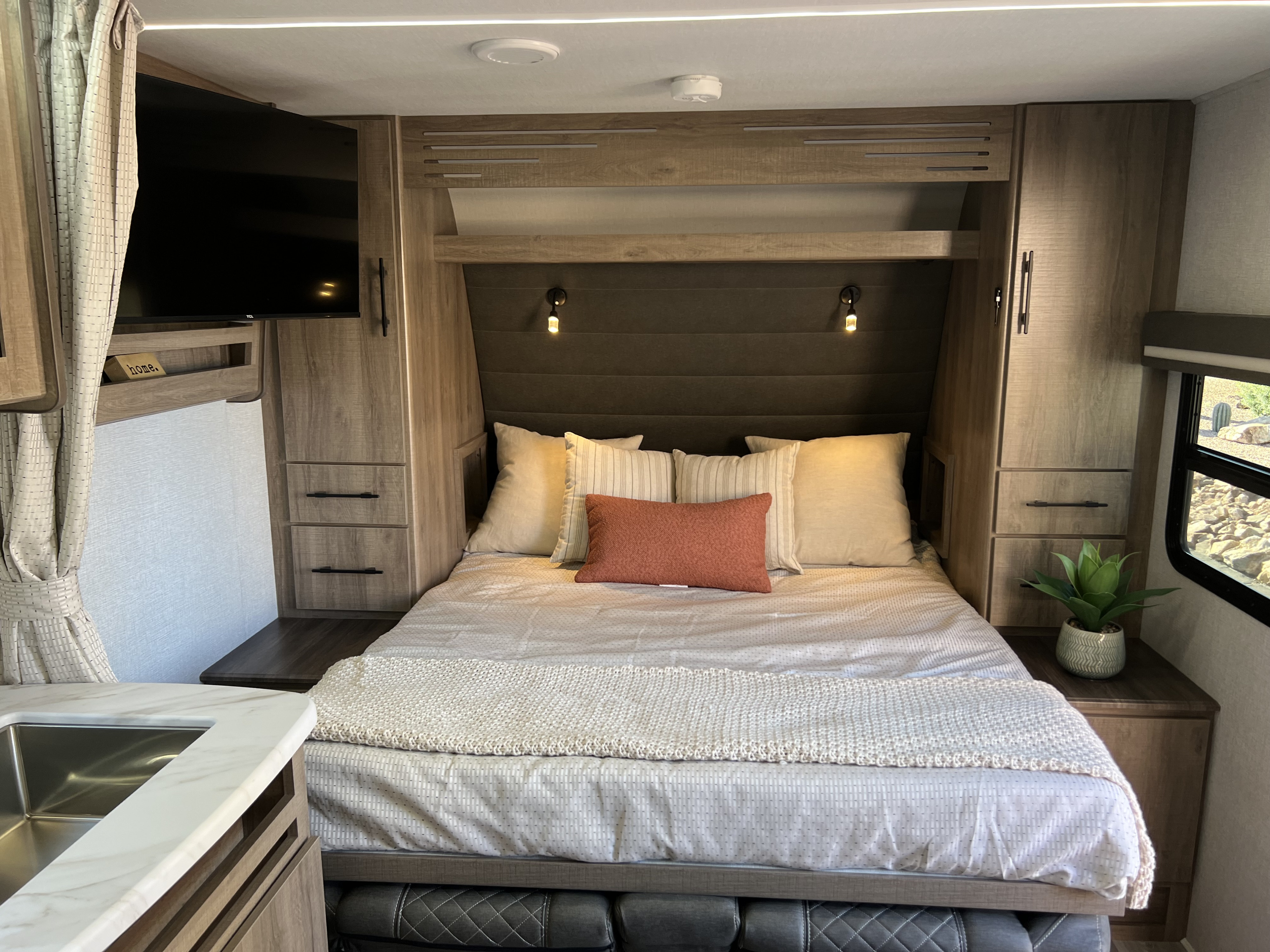Cozy RV rental bedroom with a comfortable bed, mounted TV, built-in storage, and partial kitchen view. Ideal for trips.. Grand Design Imagine XLS 2024