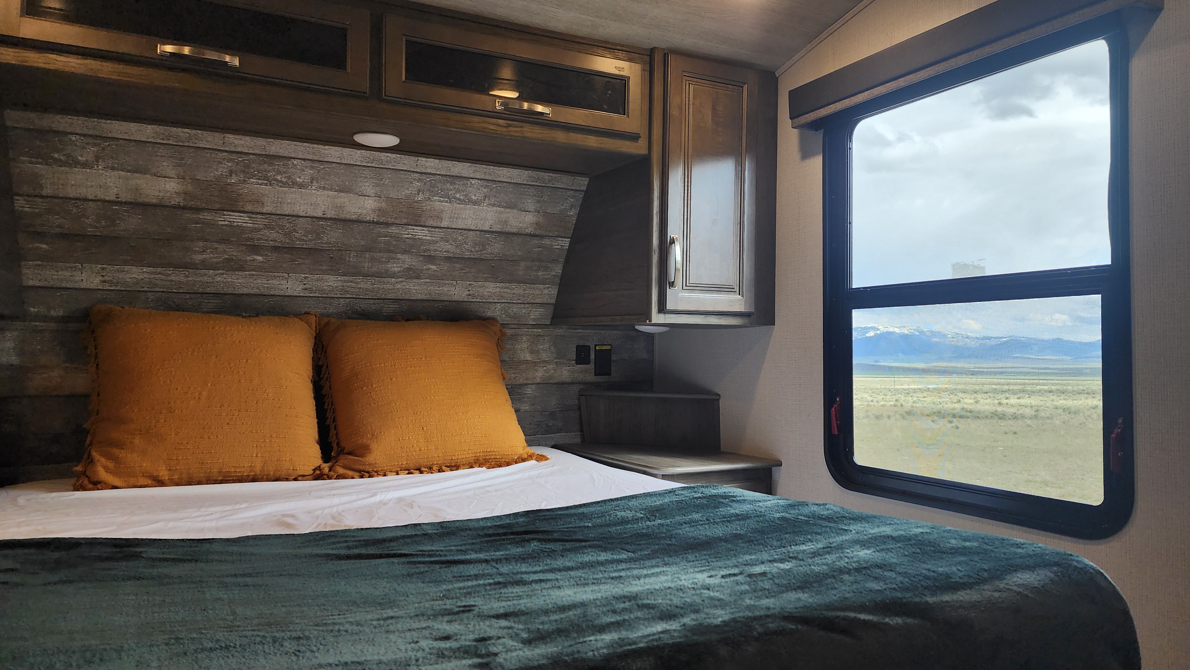 RV rental bedroom with a bed, rustic headboard, overhead cabinets, and a wide window view of distant mountains and plains.. Keystone RV Cougar 2023