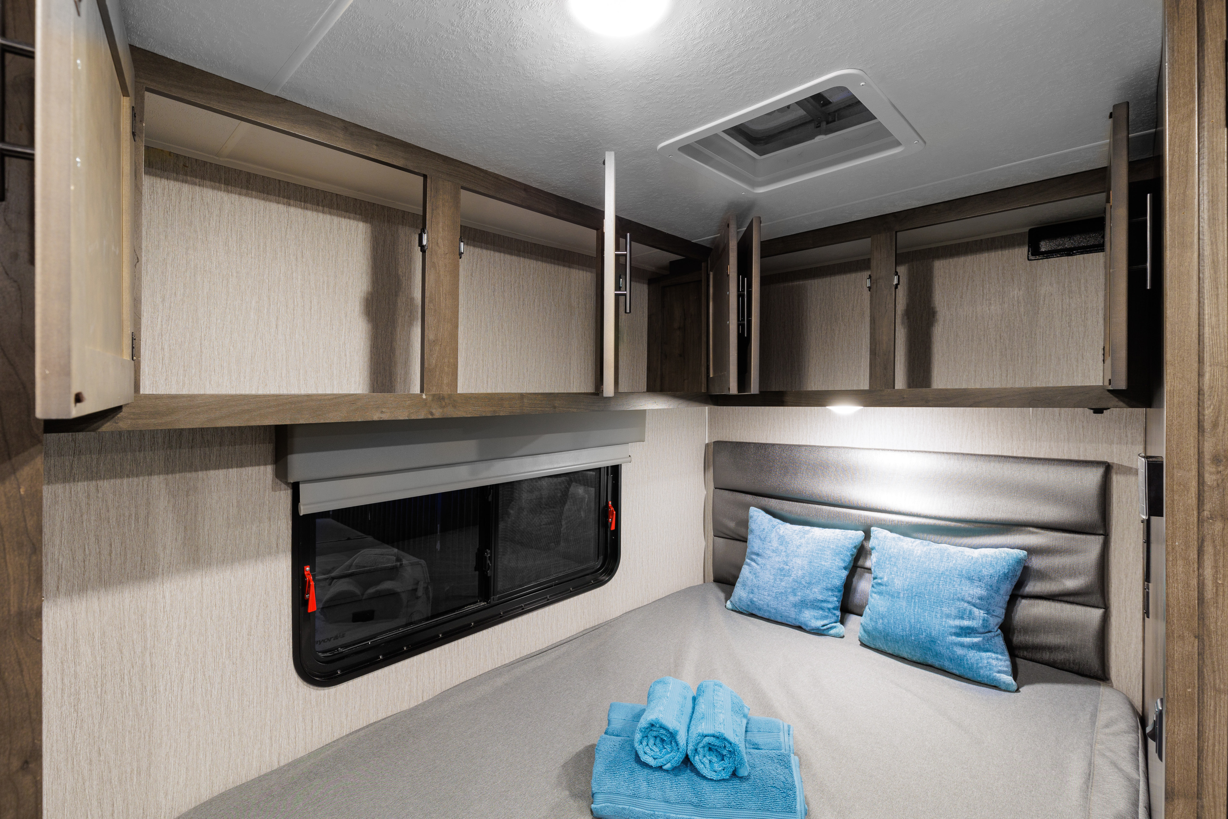 RV rental bedroom with a comfy bed, blue pillows, window, and open overhead storage cabinets, perfect for travel.. Coachmen Prism 2024