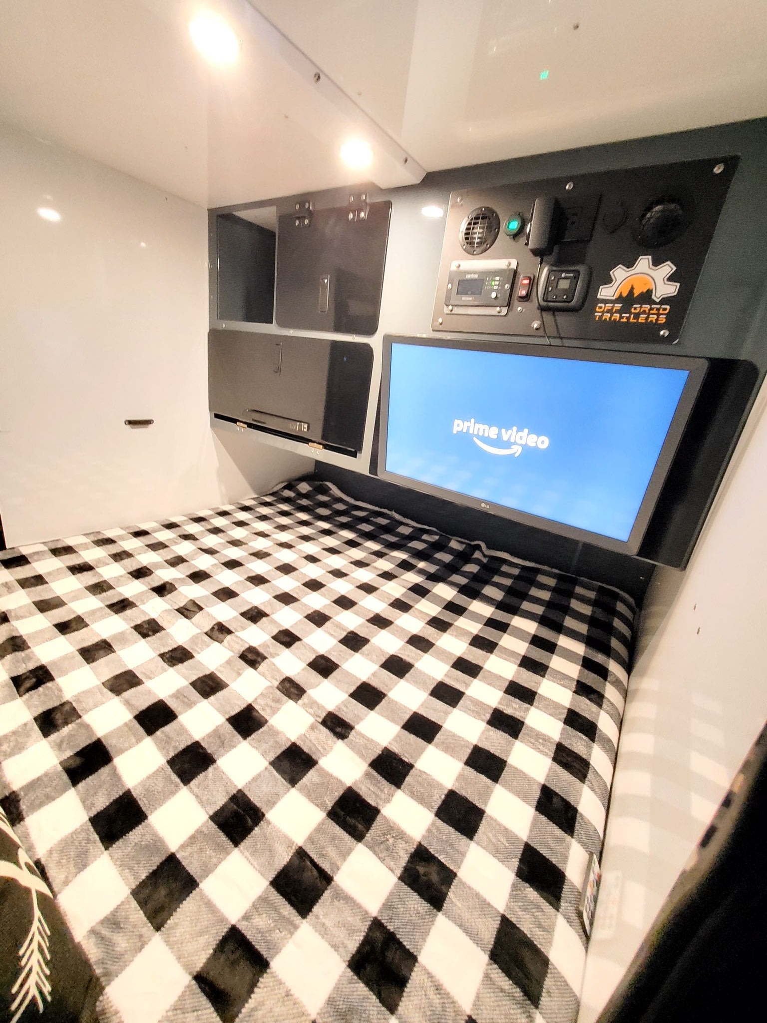Cozy RV rental interior: a comfortable bed with a checkered blanket, TV showing Prime Video, and an off-grid control panel.. Off Grid Trailers Off Grid Trailers Trailer 2021