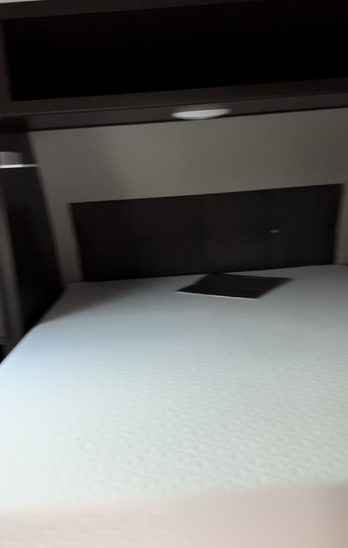 Compact RV rental bedroom featuring a white mattress, dark headboard, and overhead cabinet with a light. Ideal for travel.. Grand Design Transcend Xplor 2023