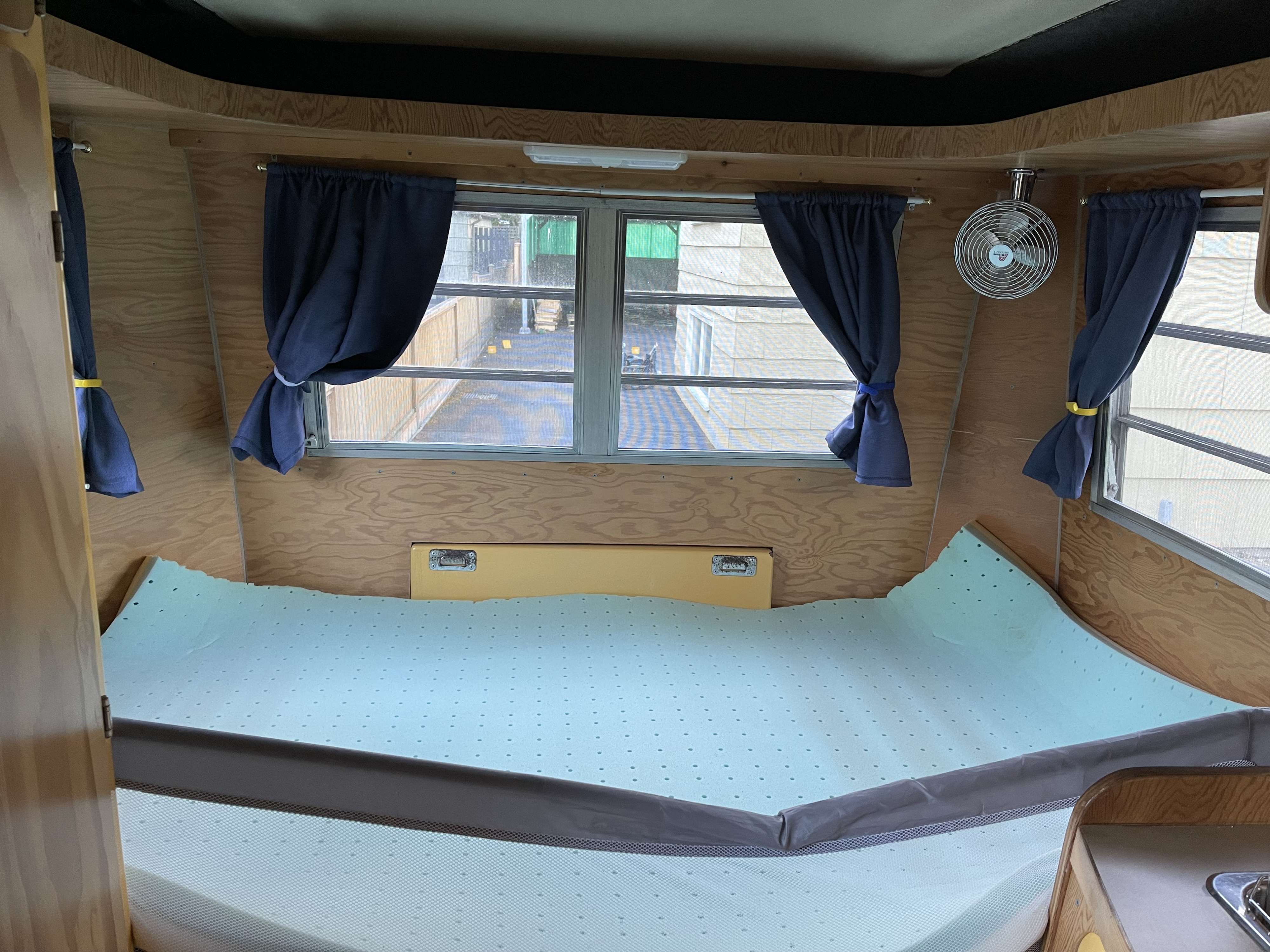 RV rental interior: A compact sleeping space with a perforated foam mattress, windows with blue curtains, and a fan.. Trillium Rv 1300 1973