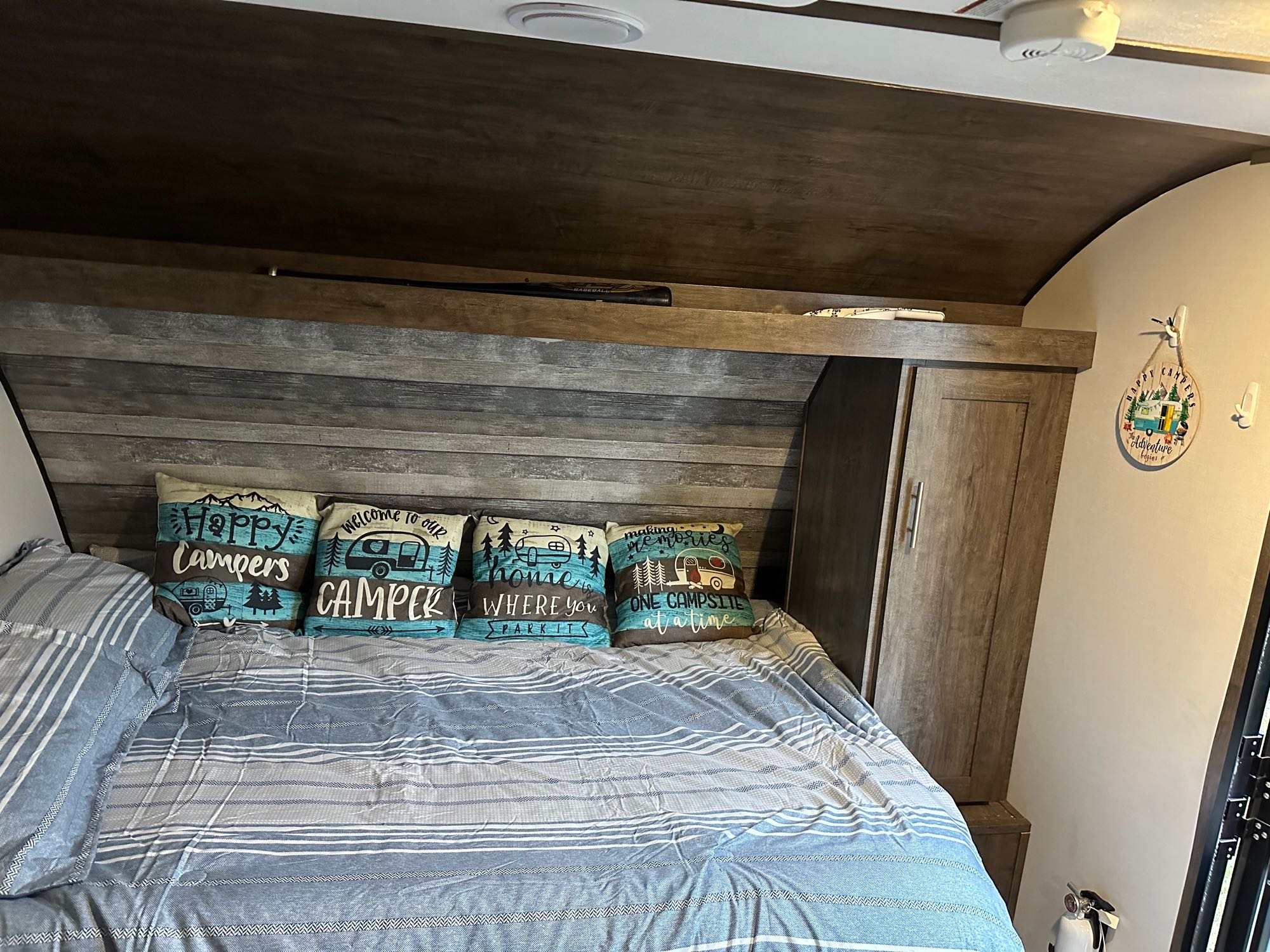 A cozy RV rental bedroom features a striped bed, four unique camping pillows, and wooden overhead storage for a comfortable stay.. Riverside RV Retro 190BH 2023