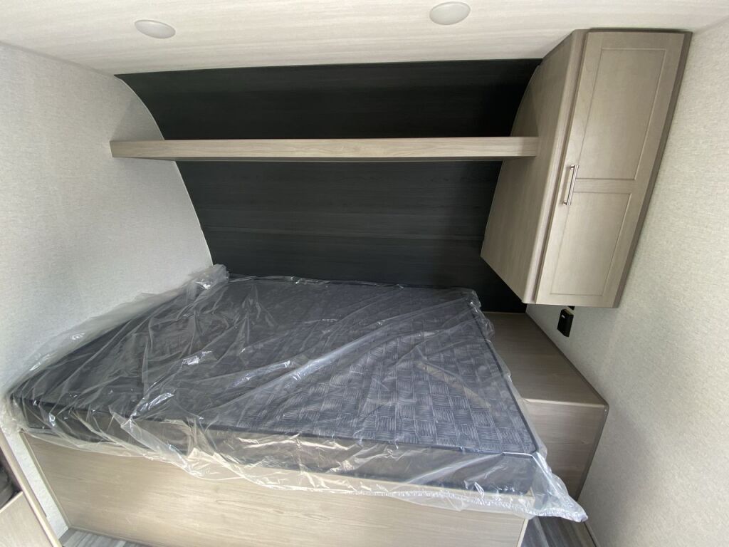 Clean RV rental bedroom features a new, plastic-covered bed, overhead shelf, and a convenient storage cabinet.. Keystone RV Springdale 2024