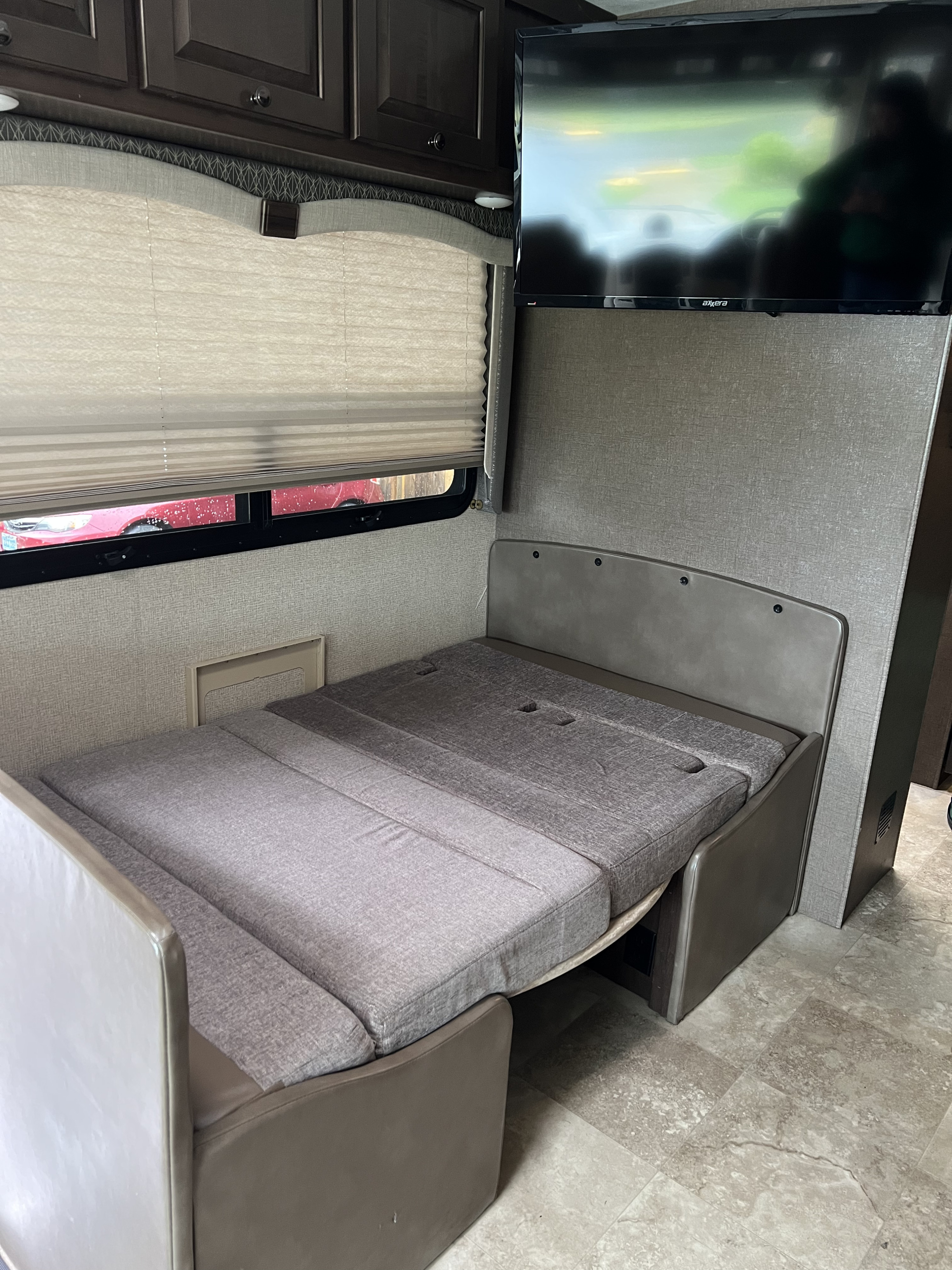 This rv rental interior features a grey upholstered bench that converts into a bed, with a wall-mounted TV, window, and overhead cabinets.. Thor Windsport 2018