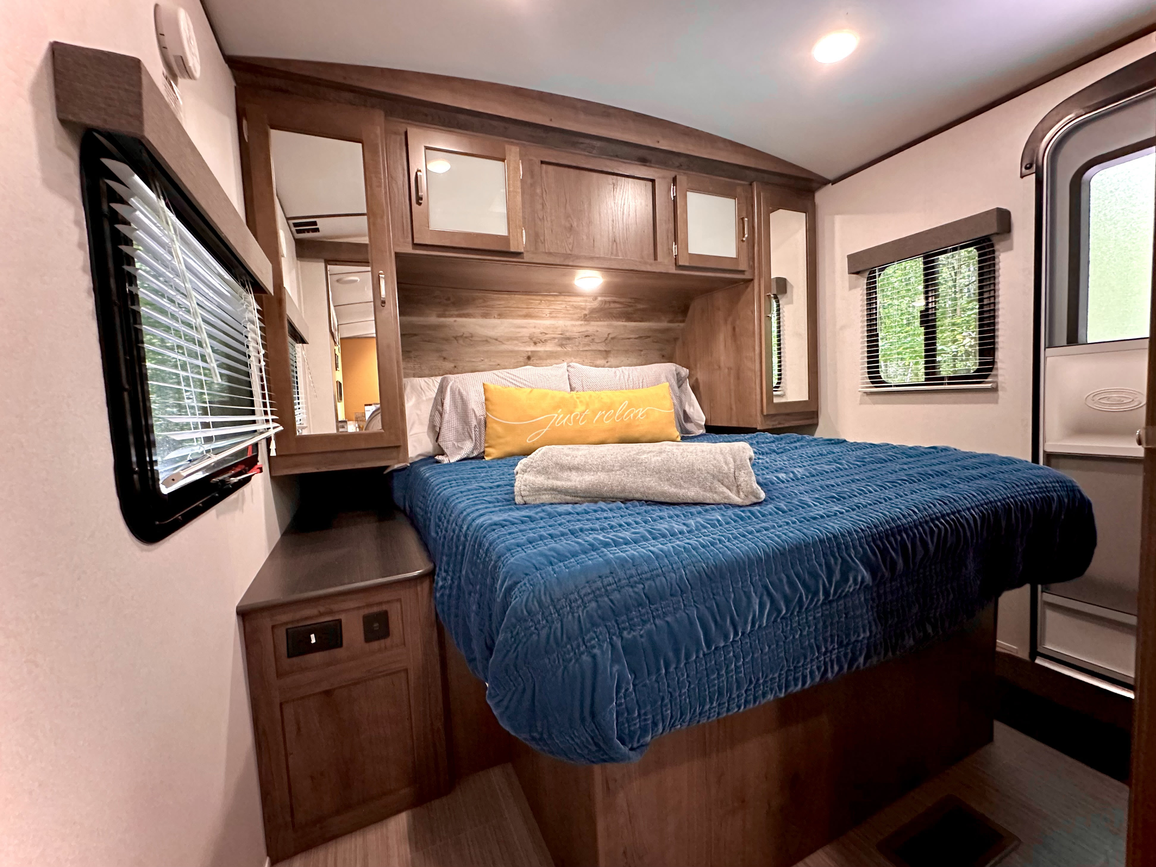 Comfortable RV rental bedroom featuring a blue quilted bed, a yellow 'just relax' pillow, and overhead wood cabinets.. Keystone RV Passport Grand Touring 2020