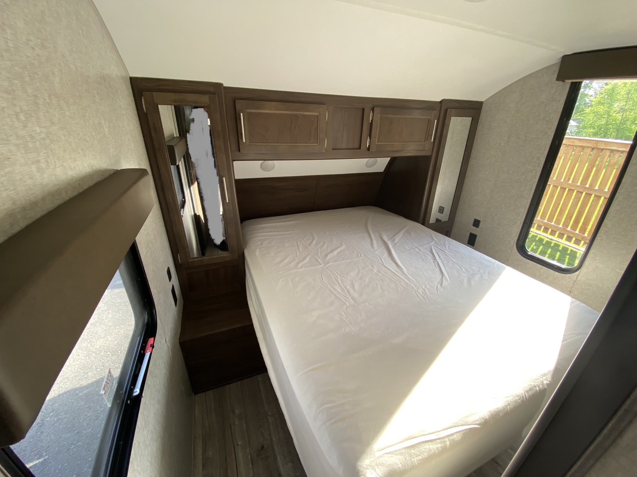 Inside an RV rental: a cozy bedroom with a white bed, dark wood cabinets, and bright windows offering views of nature and pavement.. Forest River Cherokee Grey Wolf 2021