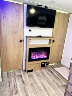 Cozy RV rental interior with a TV, an electric fireplace showing purple flames, and wood-paneled storage.. Forest River Cherokee 2024