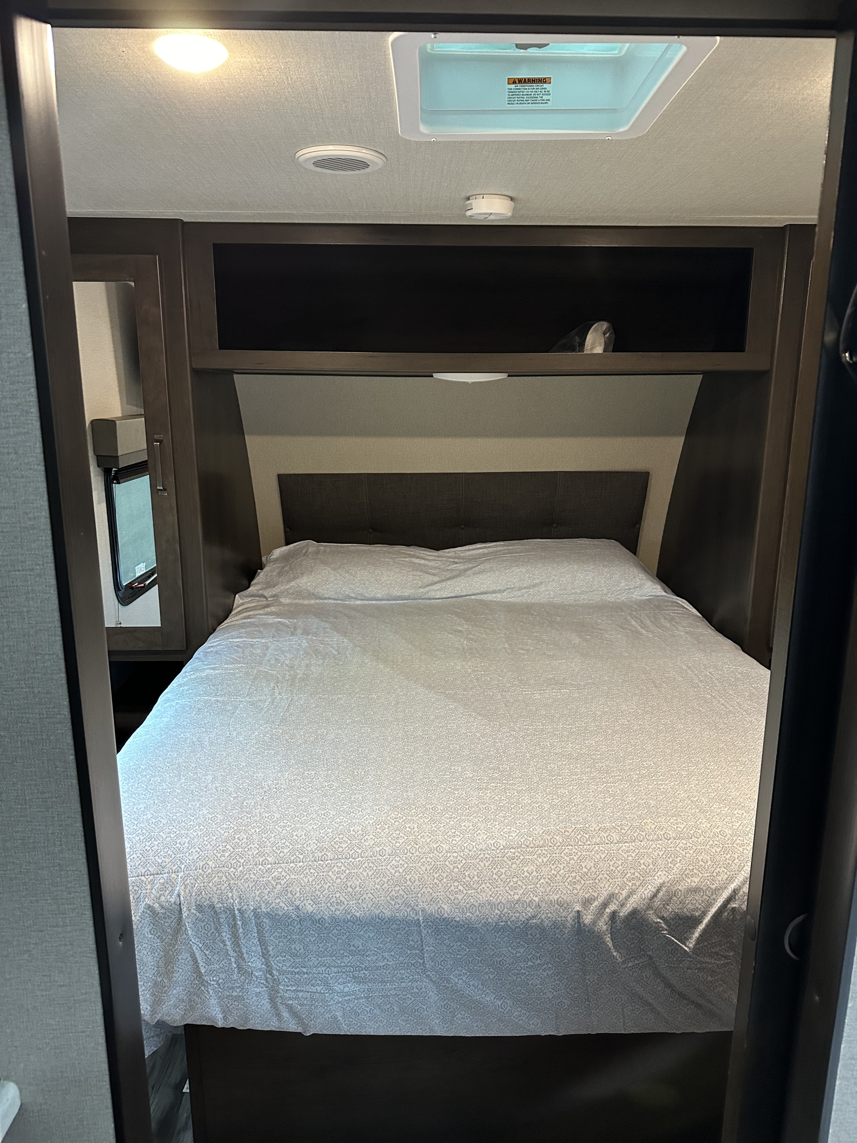 This RV rental bedroom features a comfy bed with patterned linens, overhead storage, and a side window.. Grand Design Transcend Xplor 2022