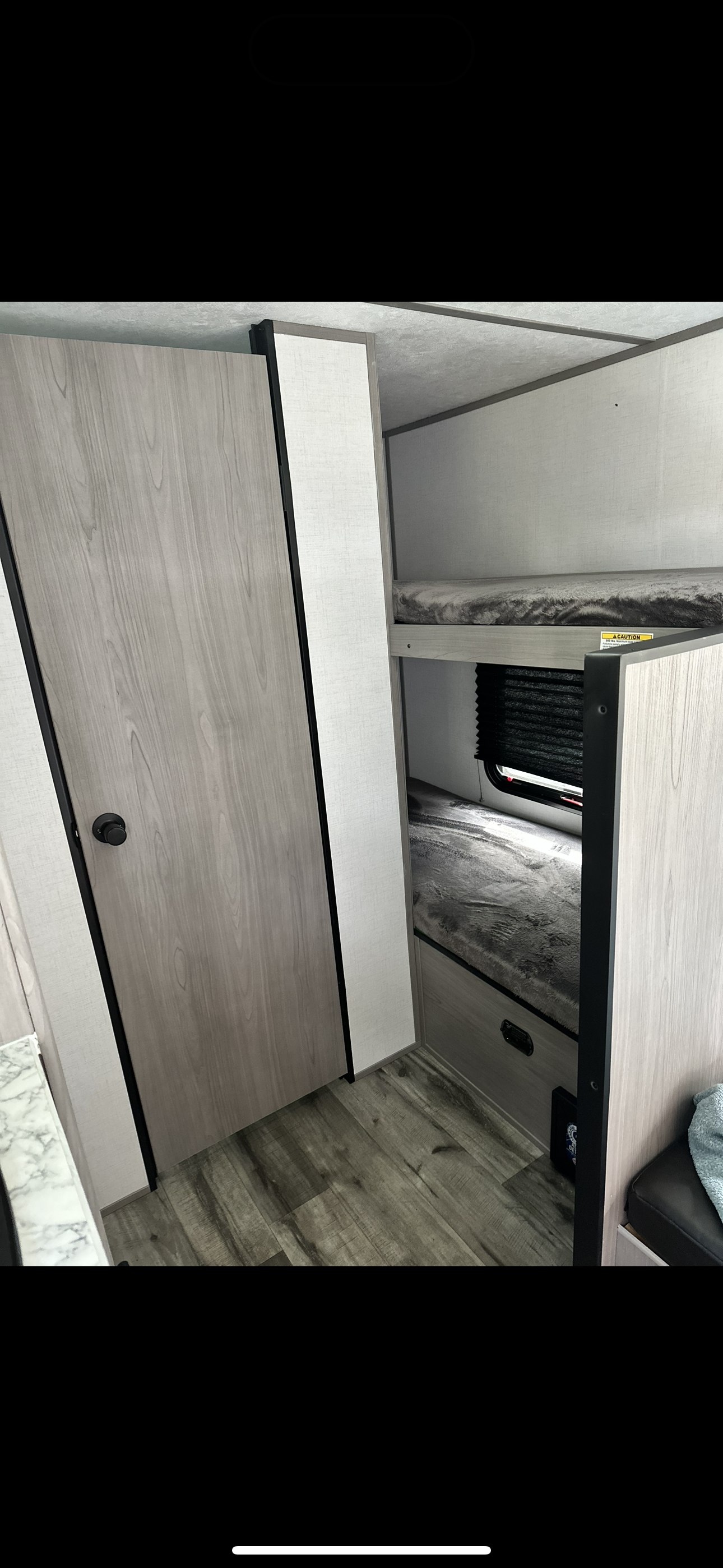 Interior view of an RV rental showing a light wood-grain door on the left, and grey bunk beds with a window blind on the right.. Coleman Lantern 17b 2024