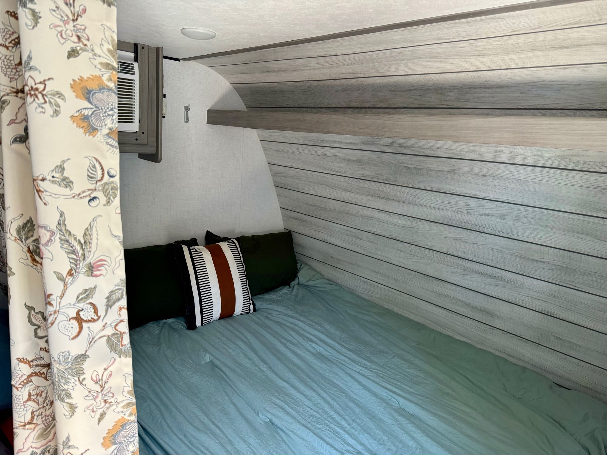 Inside this RV rental: a cozy bedroom with a bed, light blue sheets, shiplap-style walls, and an AC unit.. Coleman Lantern 17B 2024
